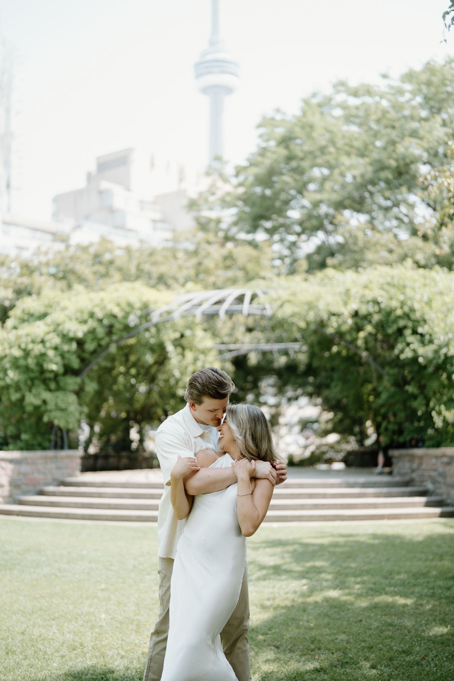 A couple embraces on a sunny day in a lush green park with trees in the background under natural lighting.