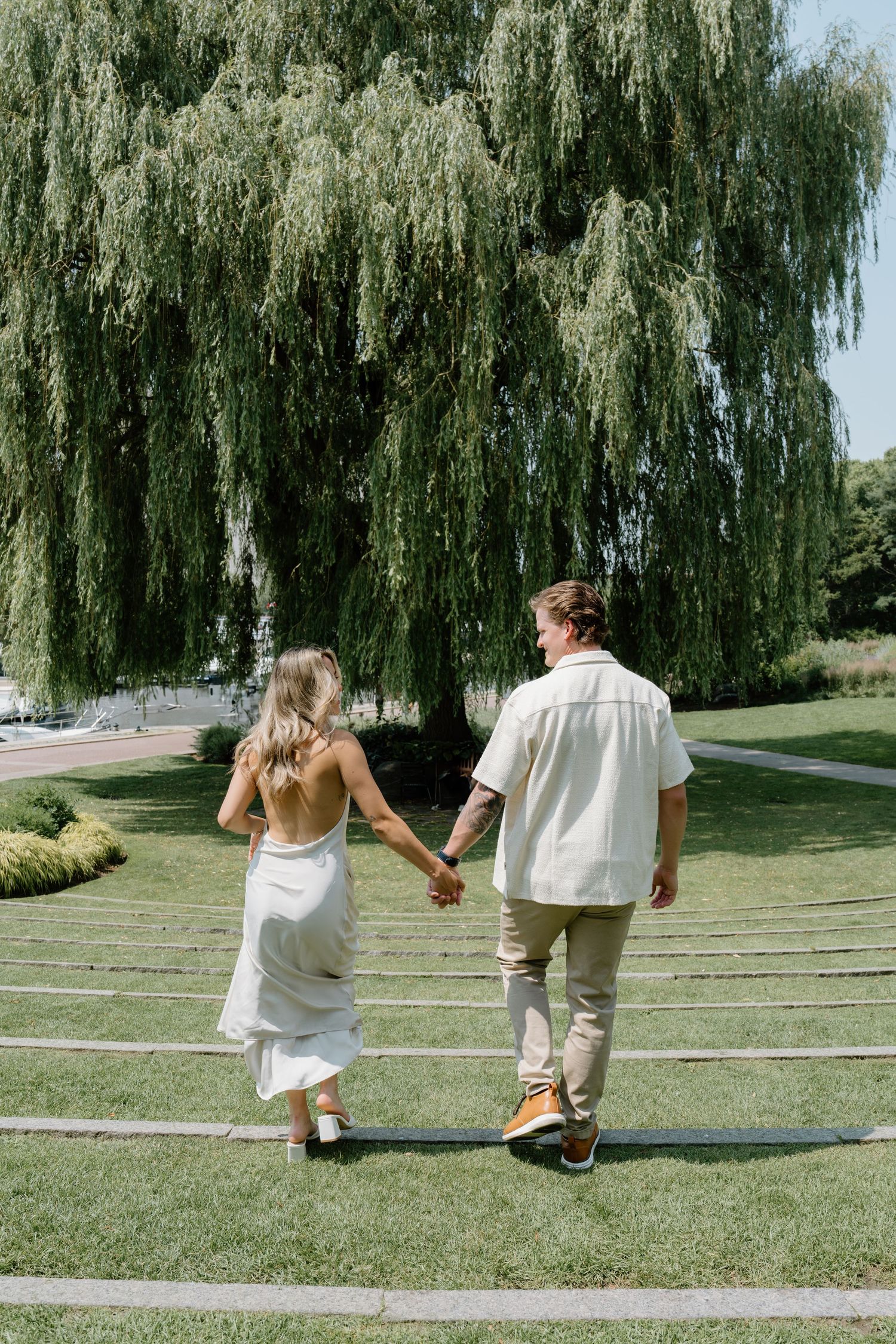 A couple holding hands walks along a curved garden path beneath a large weeping willow tree on a sunny day.
