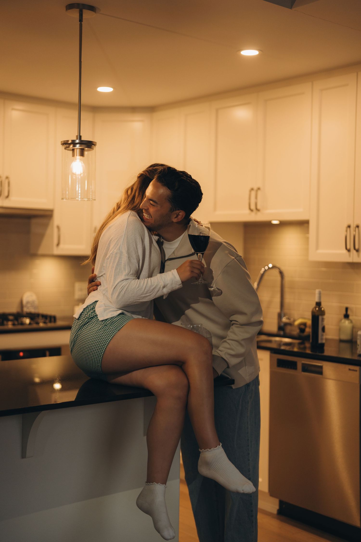 A romantic moment unfolds in a modern kitchen with warm lighting and white cabinets as a couple embraces on the countertop.