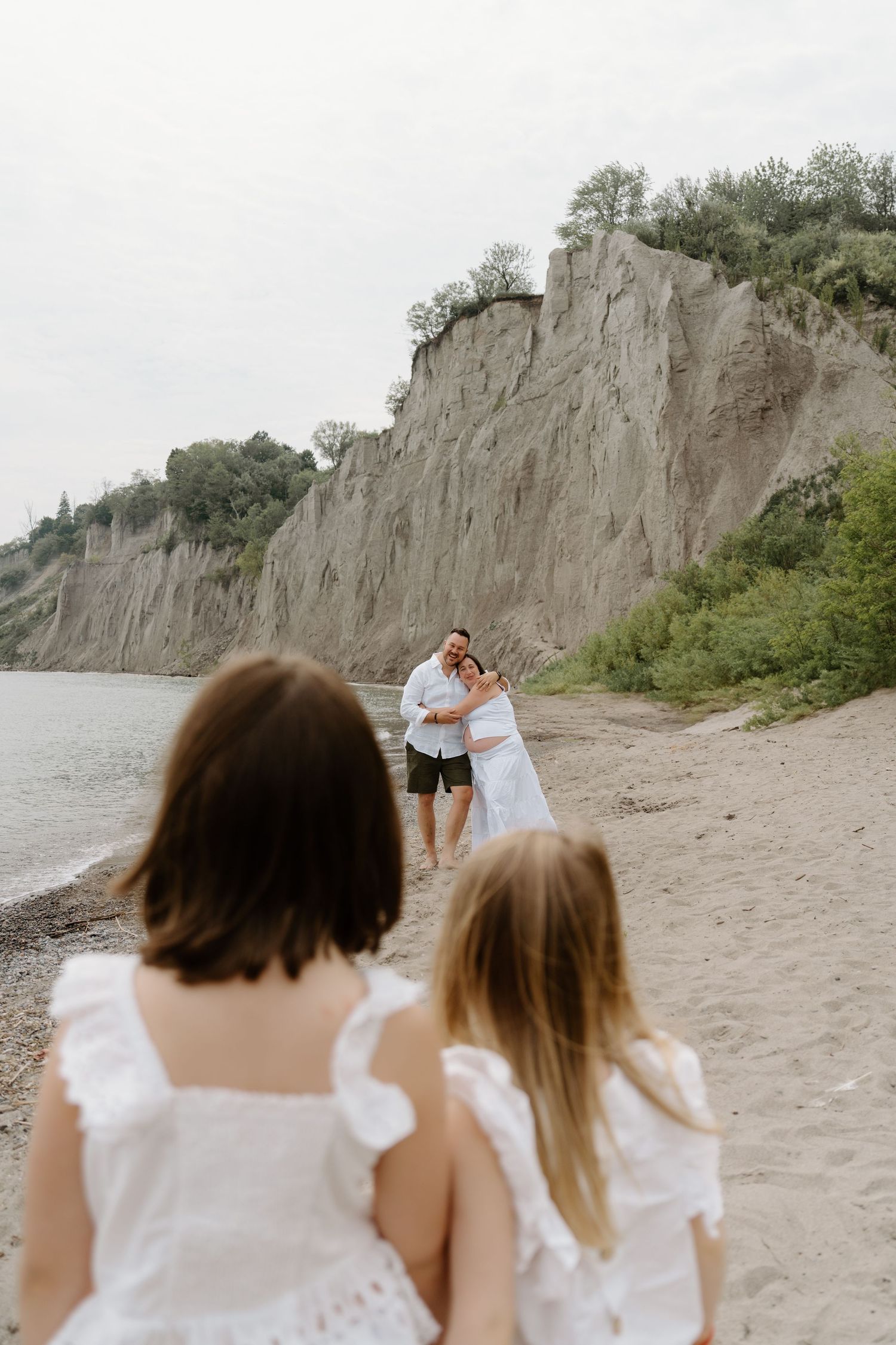 Two people in white dresses walk along a sandy beach with tall limestone cliffs in the background.