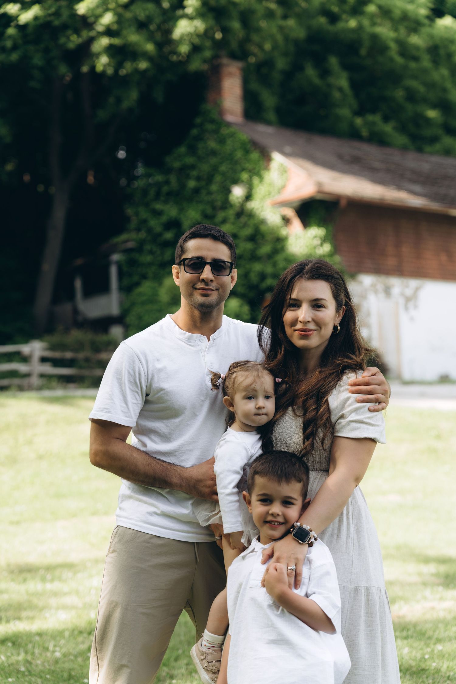 A family in coordinating white outfits poses together in front of a rustic barn on a sunny summer day.