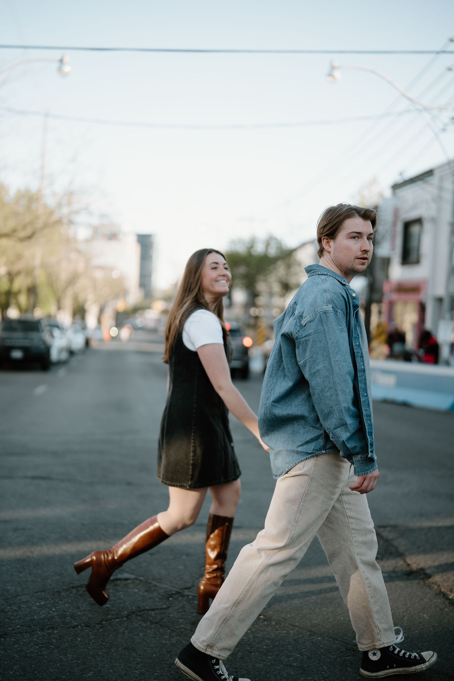 Young couple walking playfully on a city street with brown boots and denim jacket on an overcast day.