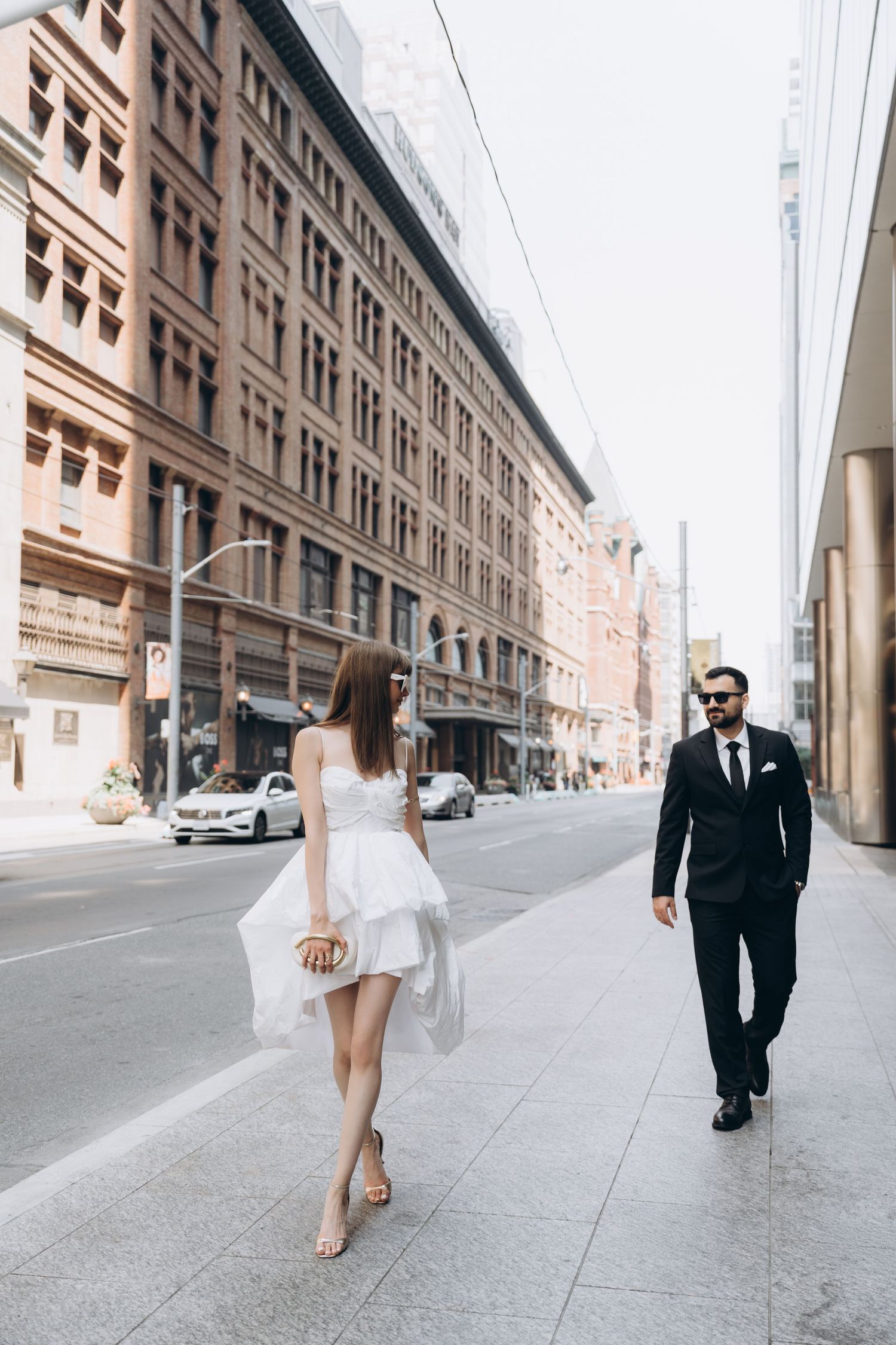 A couple in formal attire walk in opposite directions on a city sidewalk with historic buildings in the background.