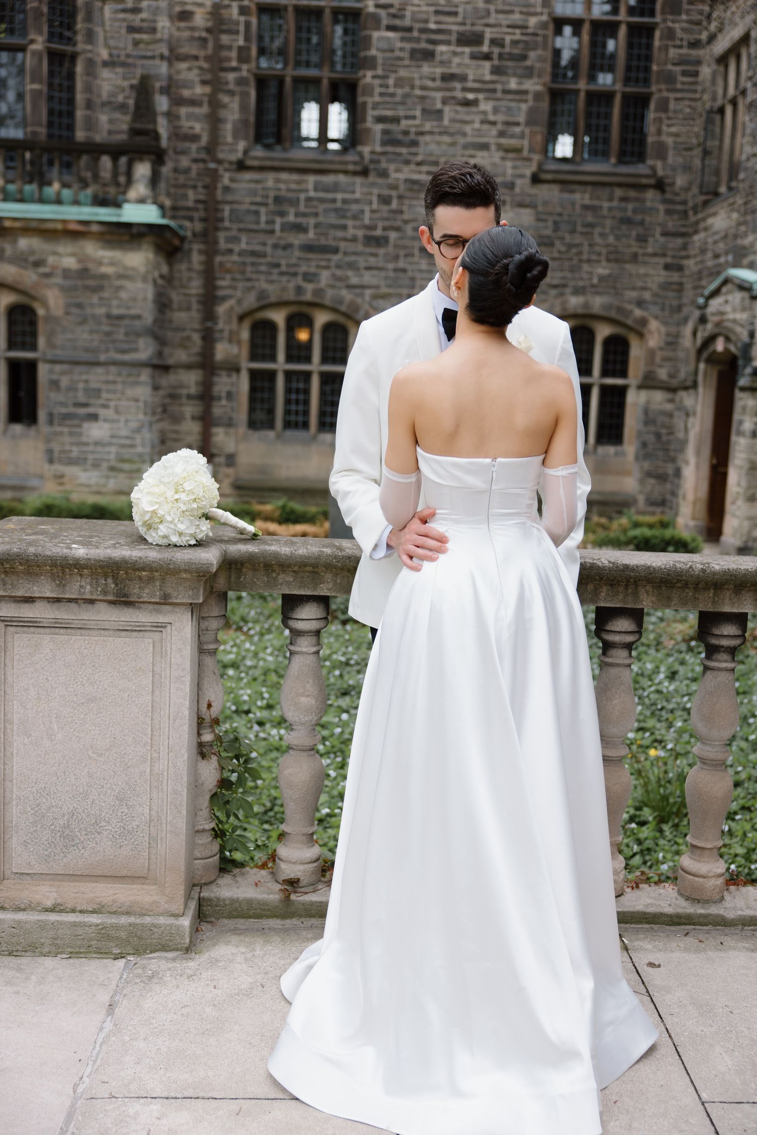 Couple in wedding attire embraces on stone balcony with white bouquet and gothic architecture in background.