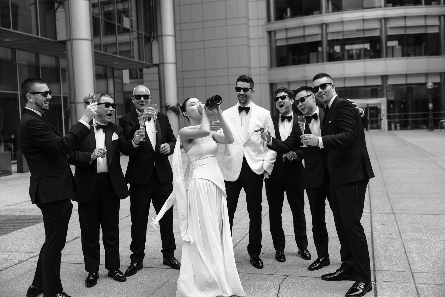 Bridal party in formal black and white attire poses together laughing outside modern building.