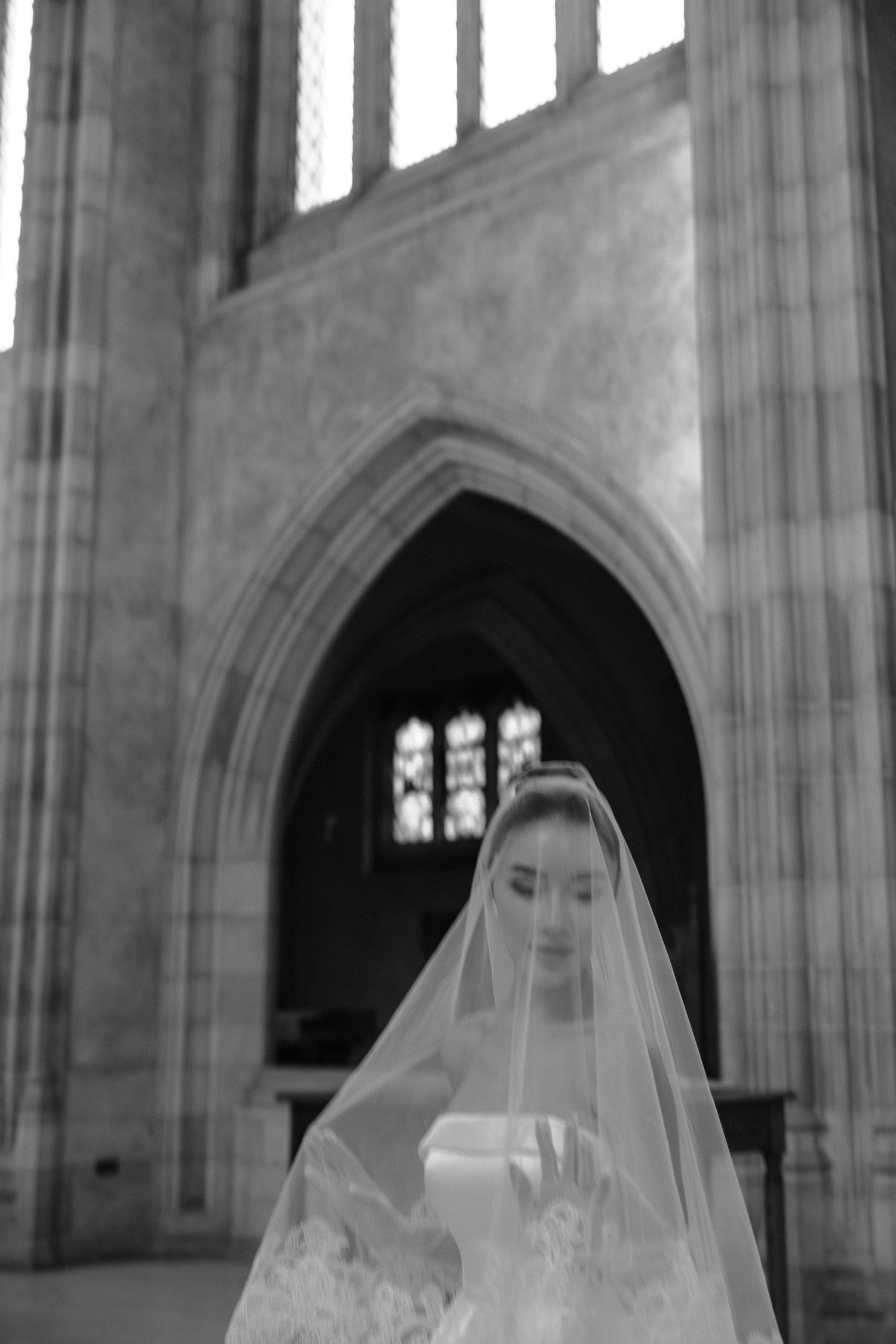 Black and white photograph of someone in an elegant bridal veil standing in a Gothic cathedral with arched windows.