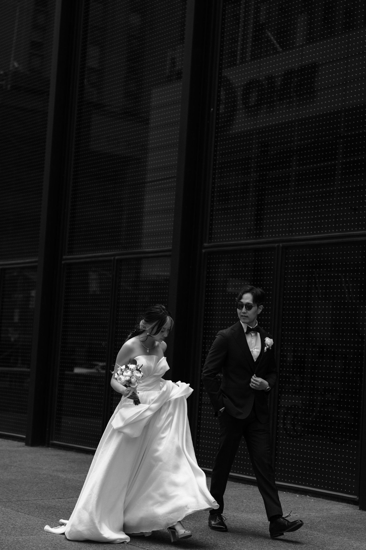 Newlyweds walk together beside a dark industrial building in dramatic black and white composition.