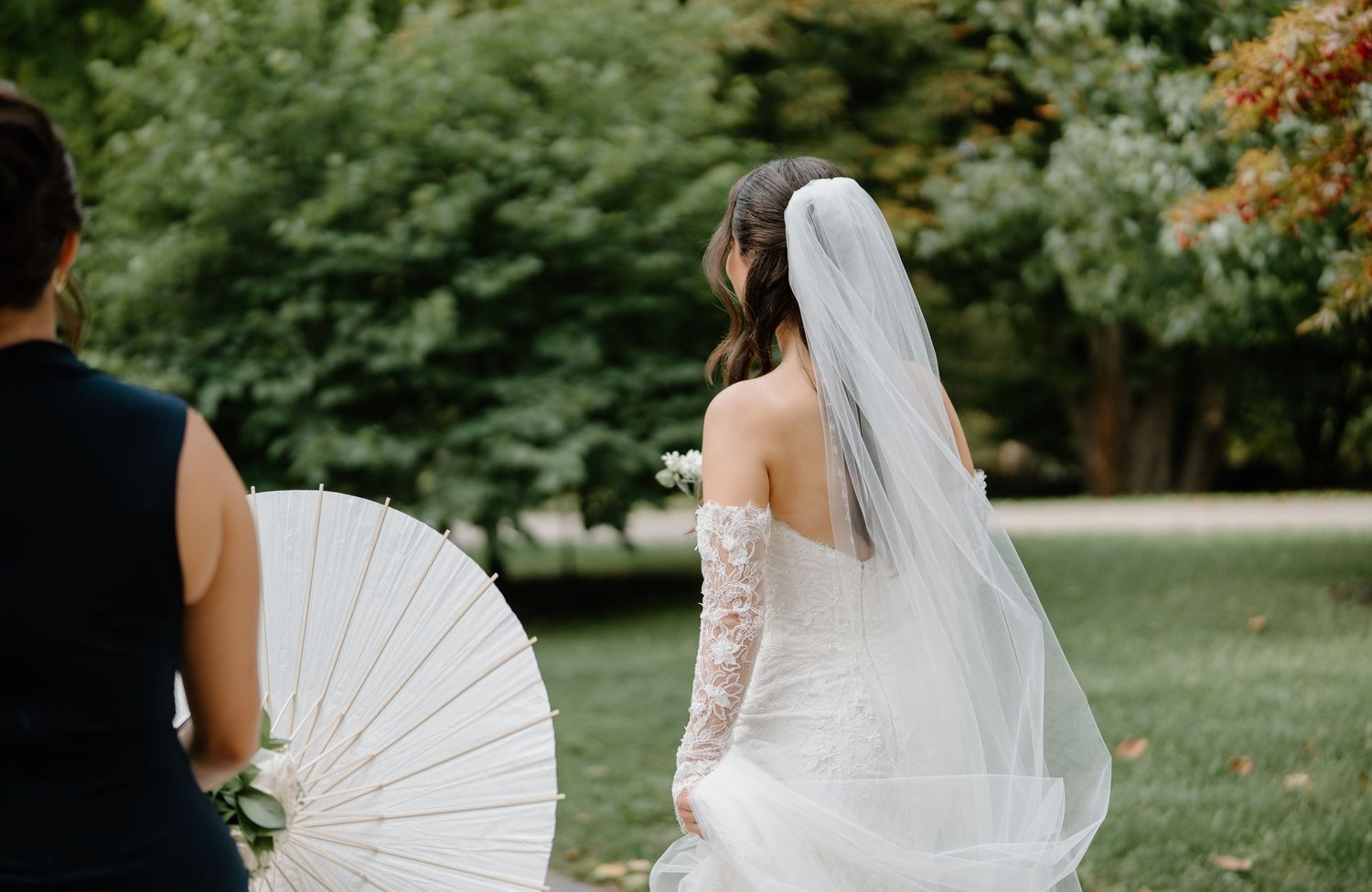 A wedding party walks through a lush garden setting while holding white parasols and flowing dresses.