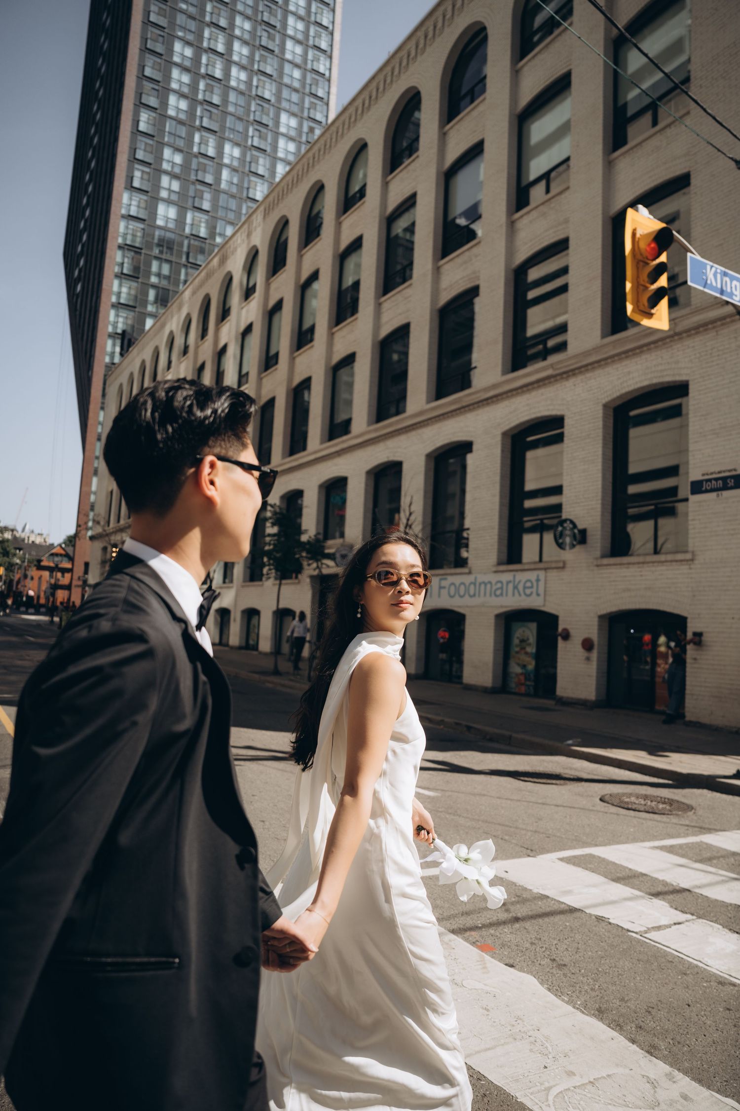 A couple in wedding attire poses dramatically on a city street with industrial buildings in the background.