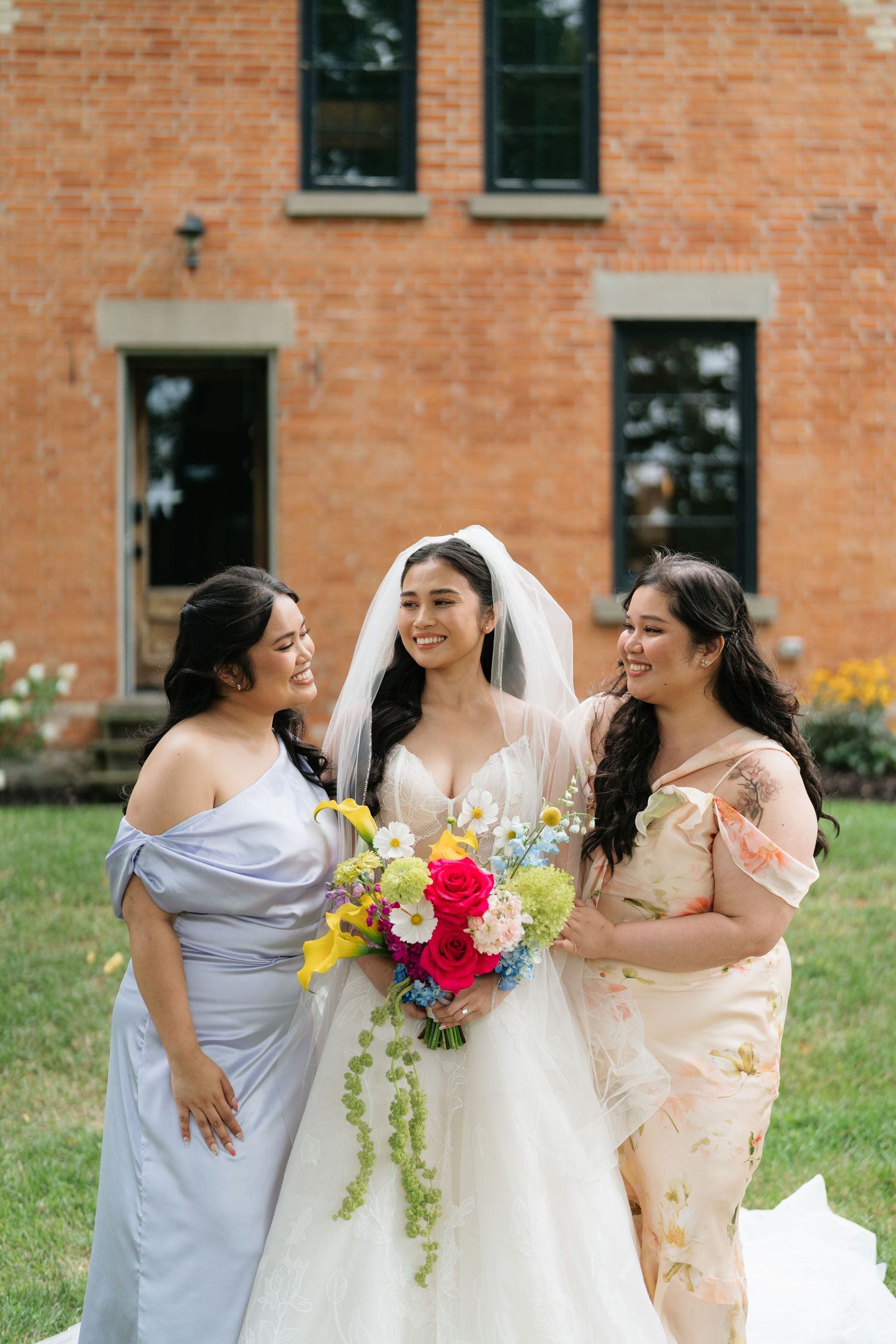 Wedding party in lavender and floral dresses share joyful moments together against brick building backdrop.