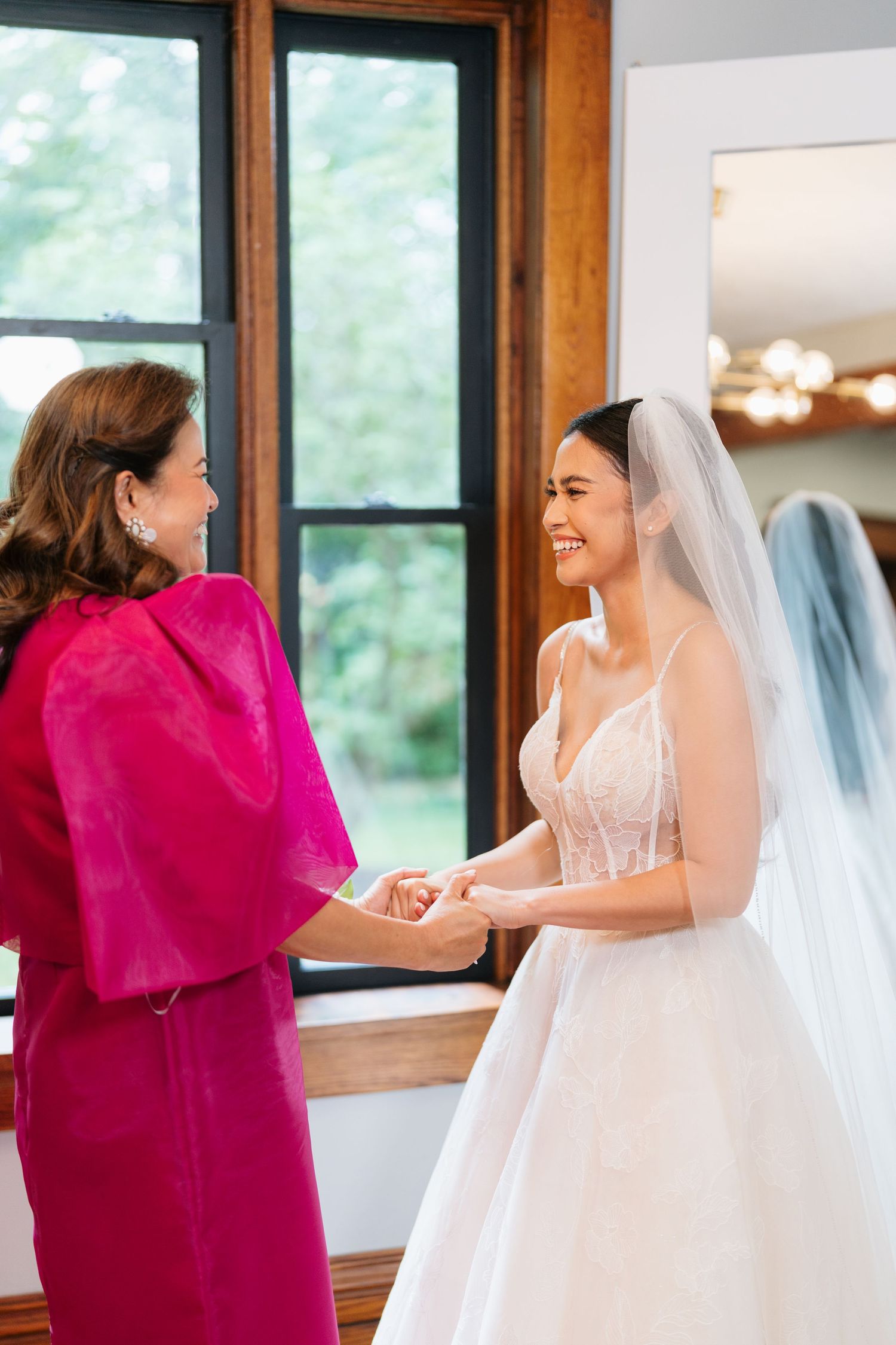 A bride in a white wedding gown shares a joyful moment with someone wearing bright magenta during wedding preparations.