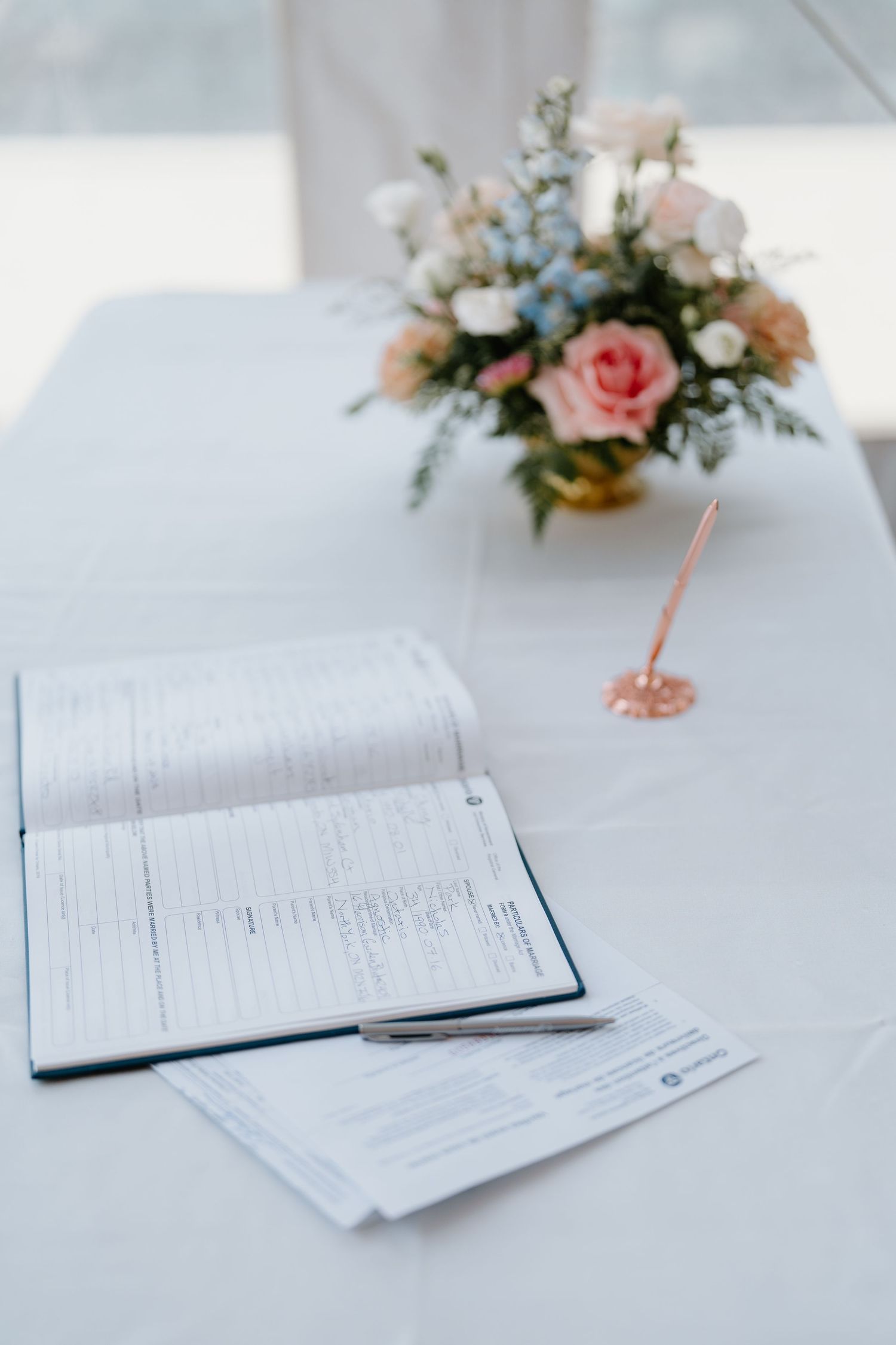 A guest book and paperwork on a white tablecloth with a soft pink and blue floral centerpiece arrangement.