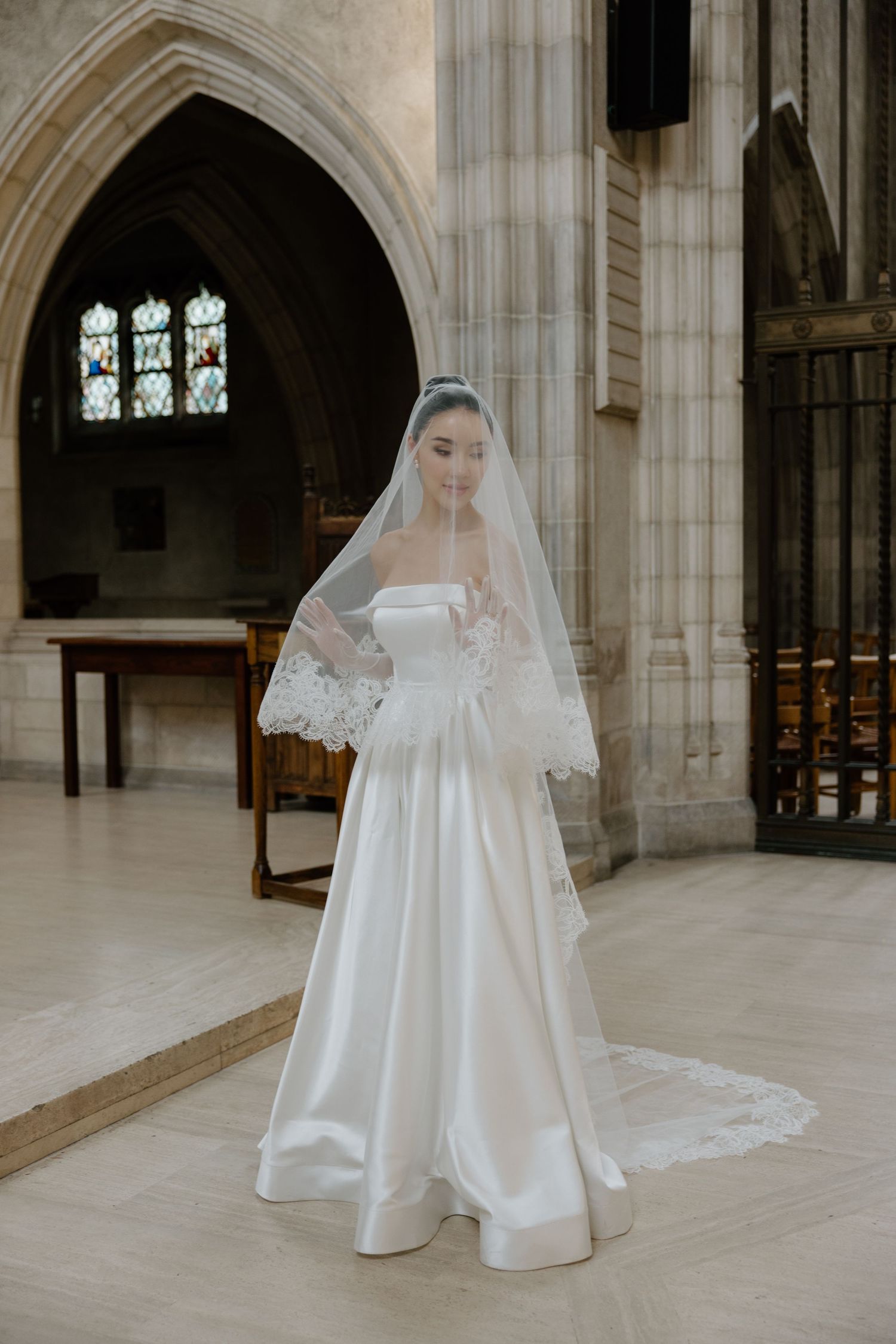 Traditional white wedding gown and lace veil photographed in a gothic church interior with arched windows and stone columns.