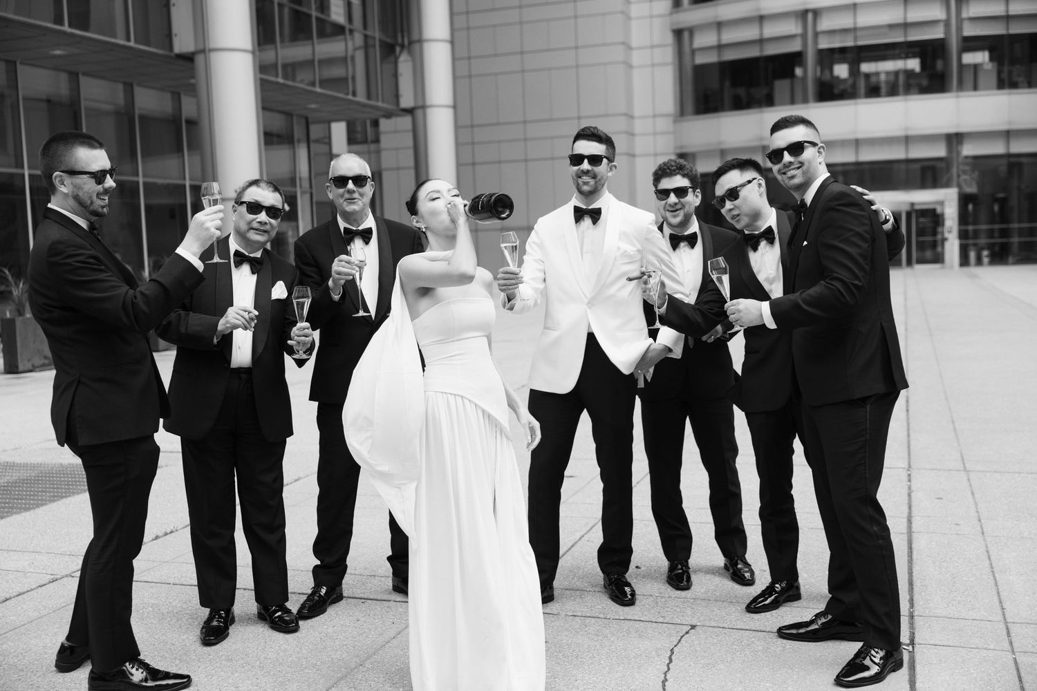 A wedding party in formal black and white attire poses together outdoors at a modern building.