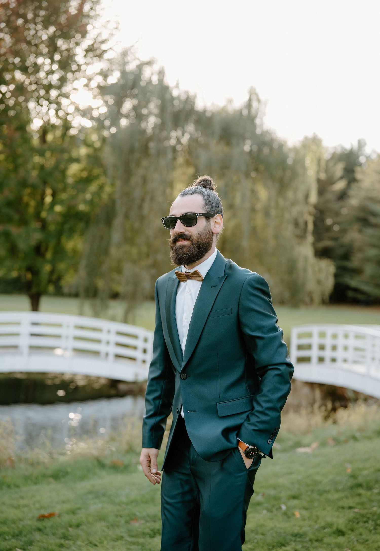 Stylish groom in teal suit and wooden bowtie stands near white fence in garden setting.