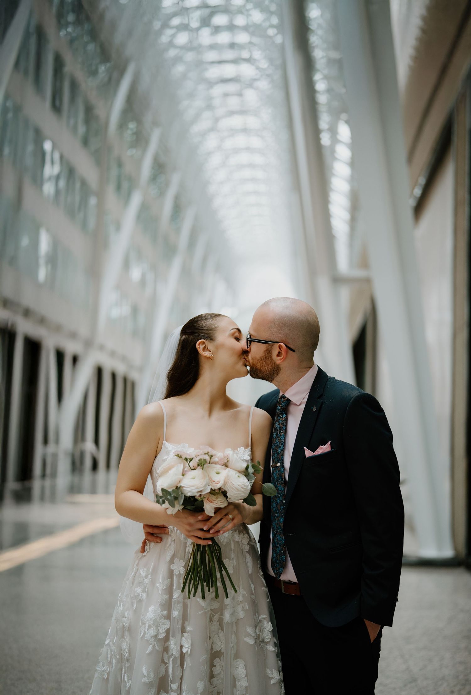 Elegant couple shares romantic kisses in a bright architectural corridor with glowing natural light and white arches.