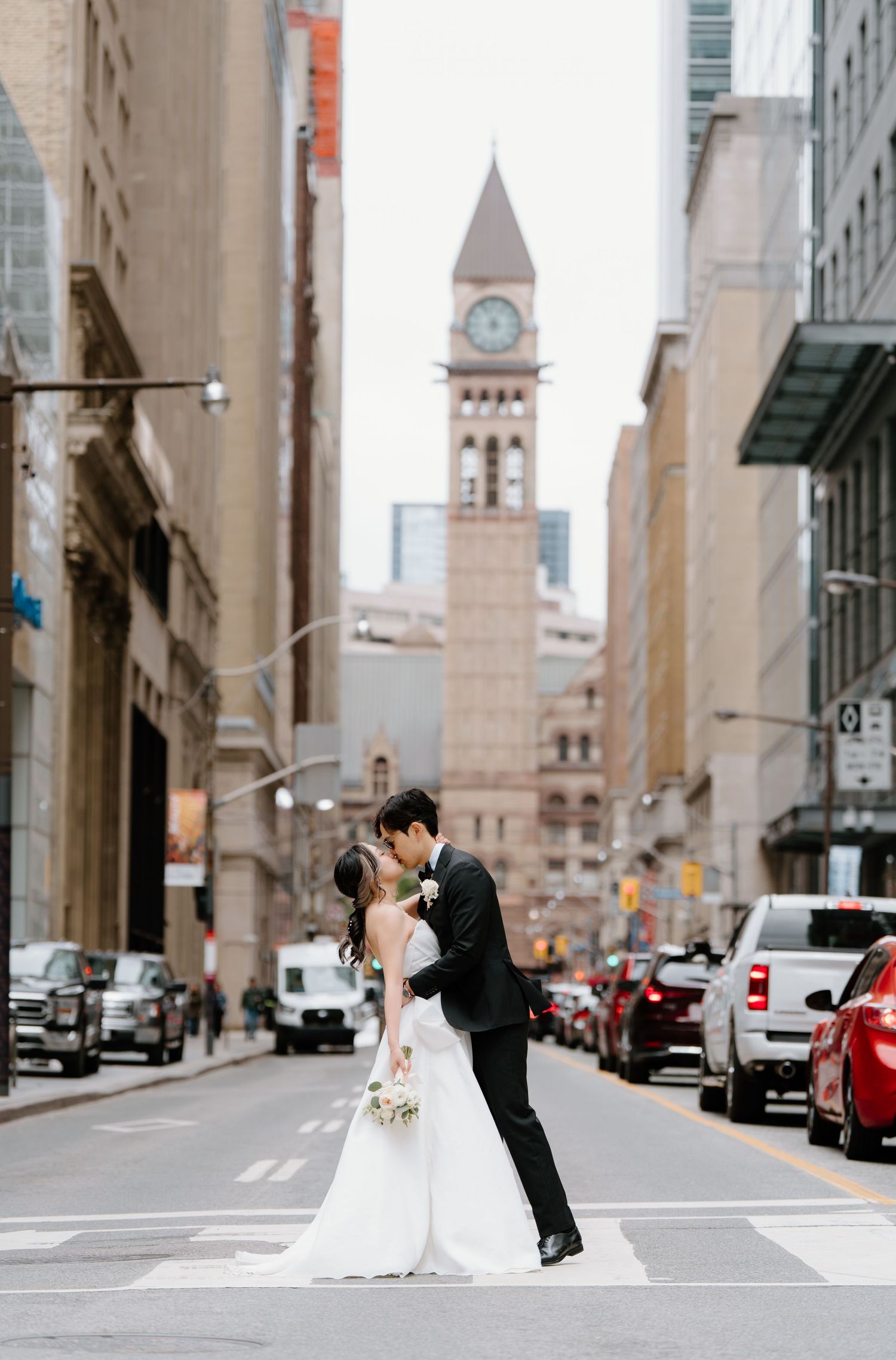 Newlyweds share romantic kiss in middle of Toronto street with historic Old City Hall clock tower in background.