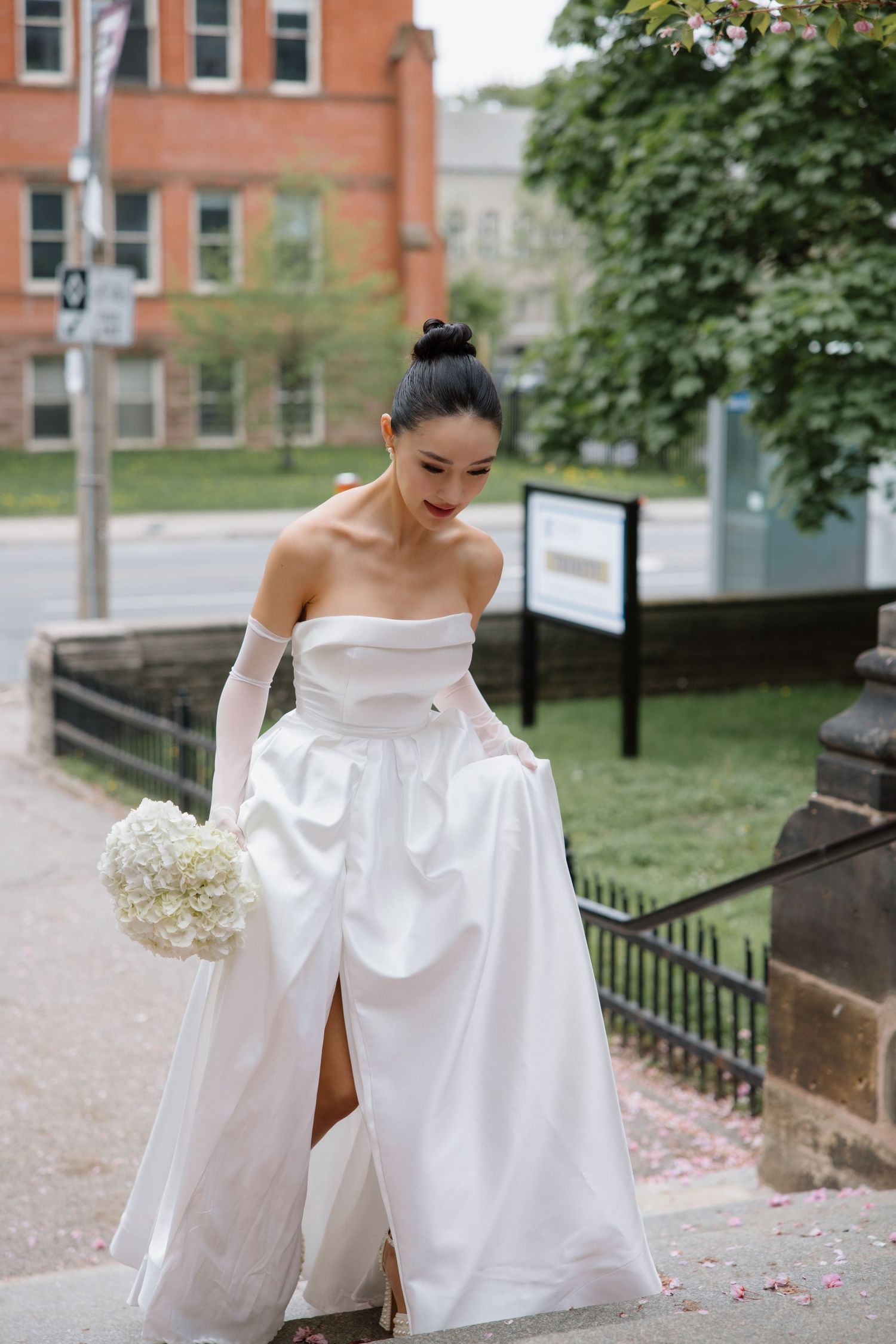 A bride in an elegant off-shoulder white wedding gown holds a bouquet while walking up stone steps near a brick building.