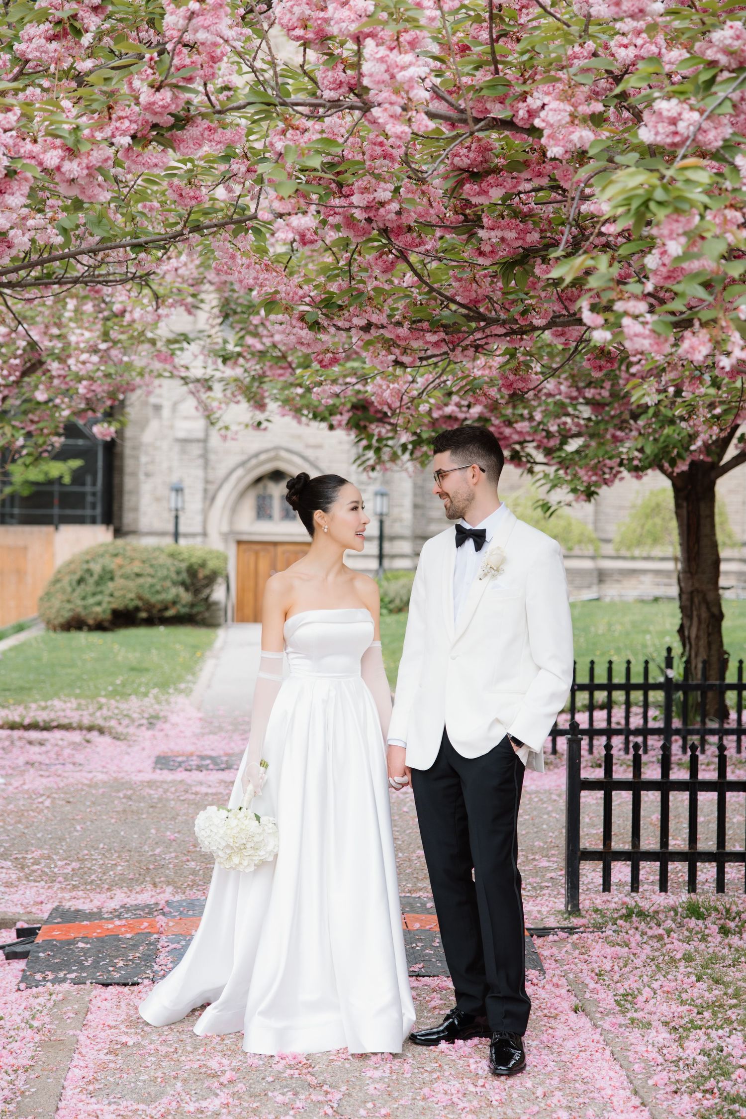 A couple stands beneath a blooming cherry blossom tree in a garden setting with a church facade in the background.