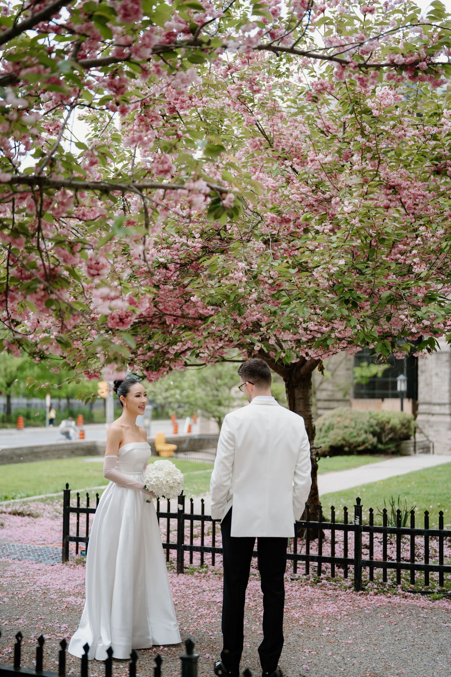 A couple stands under blooming cherry blossom trees with a white dress and formal attire during a spring wedding.
