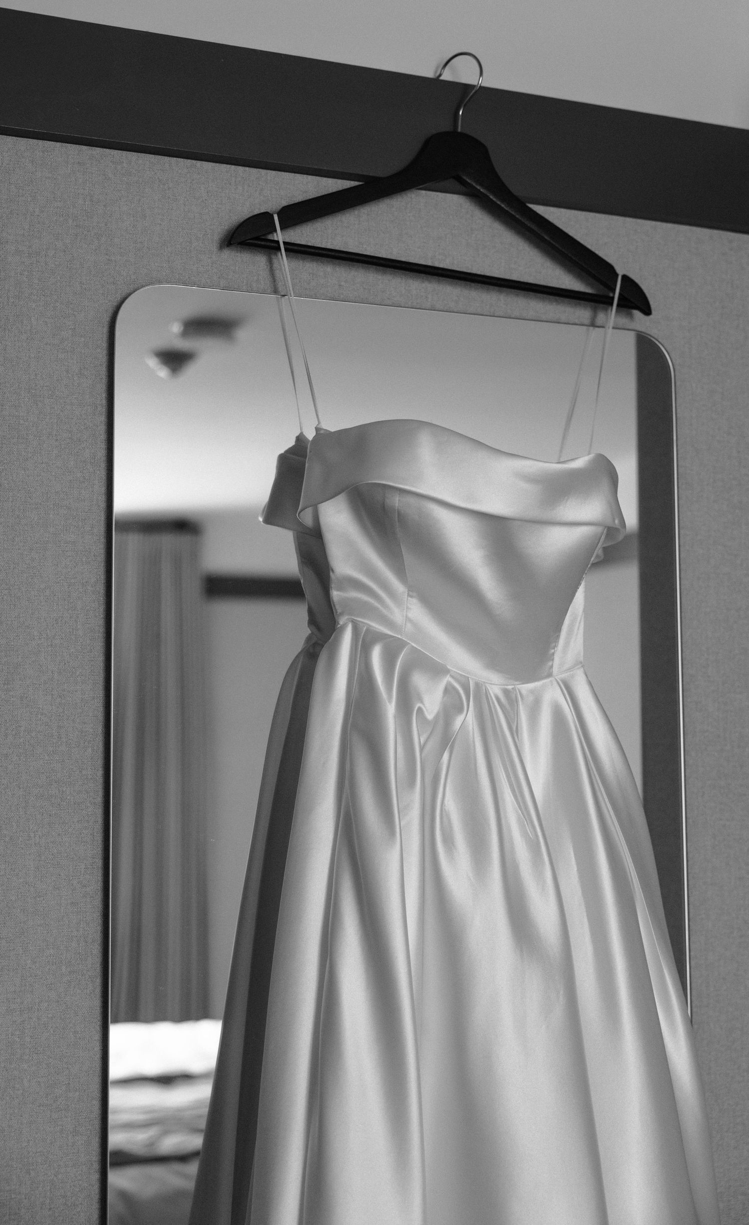 A classic white satin wedding dress hangs on a black hanger against a gray wall, displaying its elegant off-shoulder design.