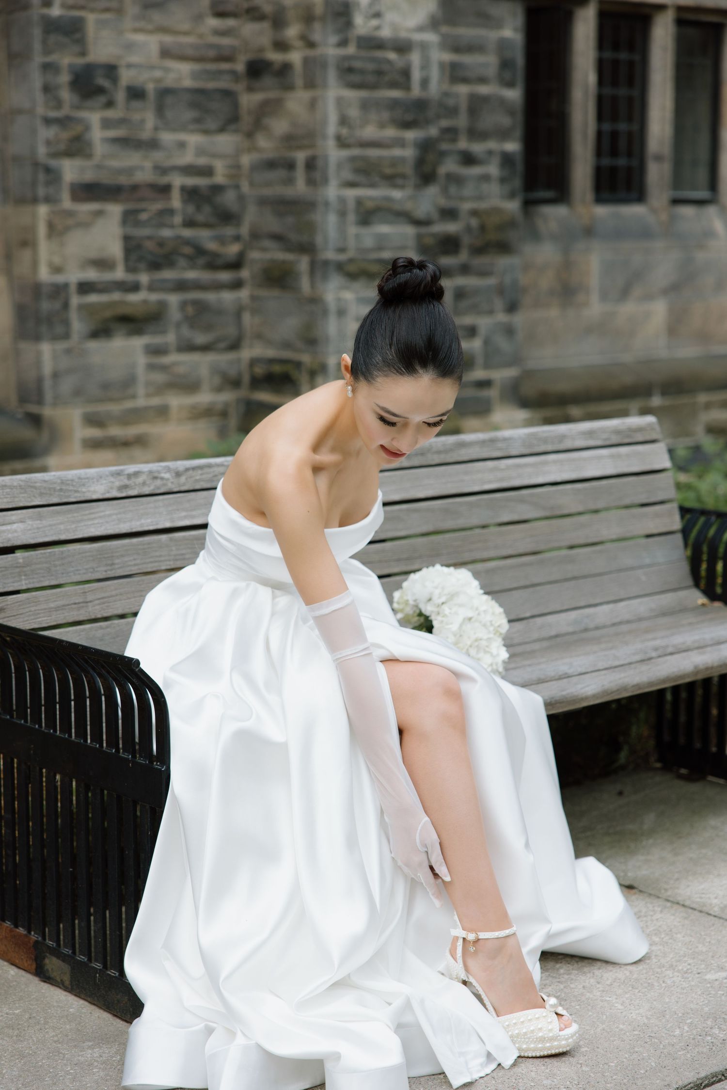 A bride in a flowing white dress adjusts her elegant white shoes while sitting on a stone bench outside a church.