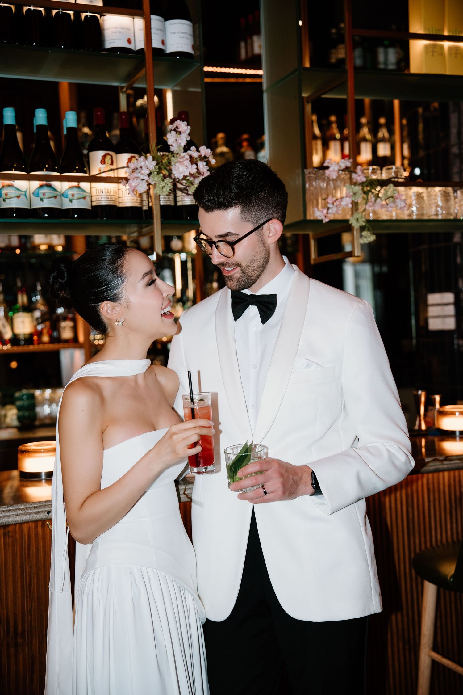 A couple in formal wedding attire share drinks at an elegant bar with shelves of bottles in the background.