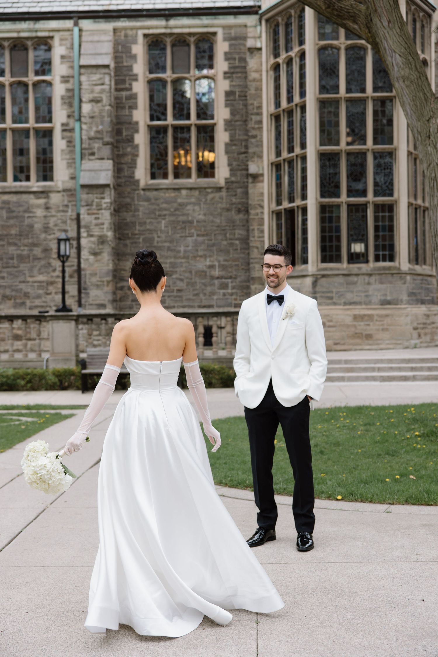 A bride in a strapless white gown stands on a path facing her partner in front of a historic stone building with gothic windows.
