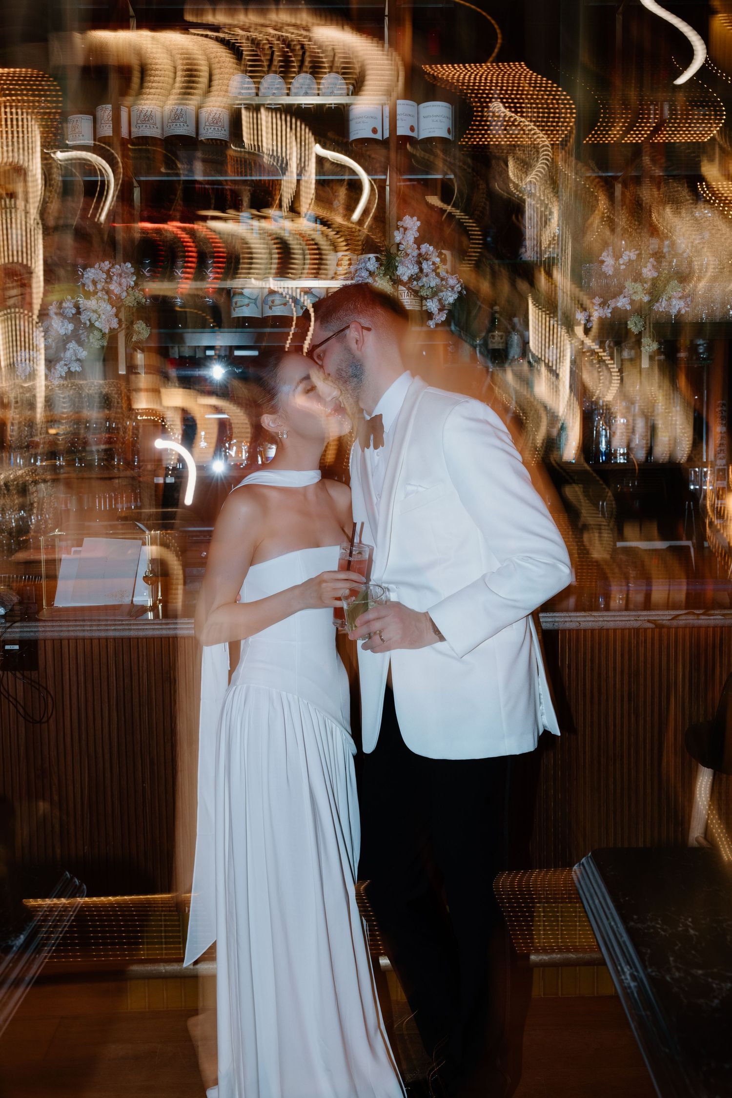A couple dances in formal attire at a venue with wine glasses hanging overhead creating a romantic ambiance.
