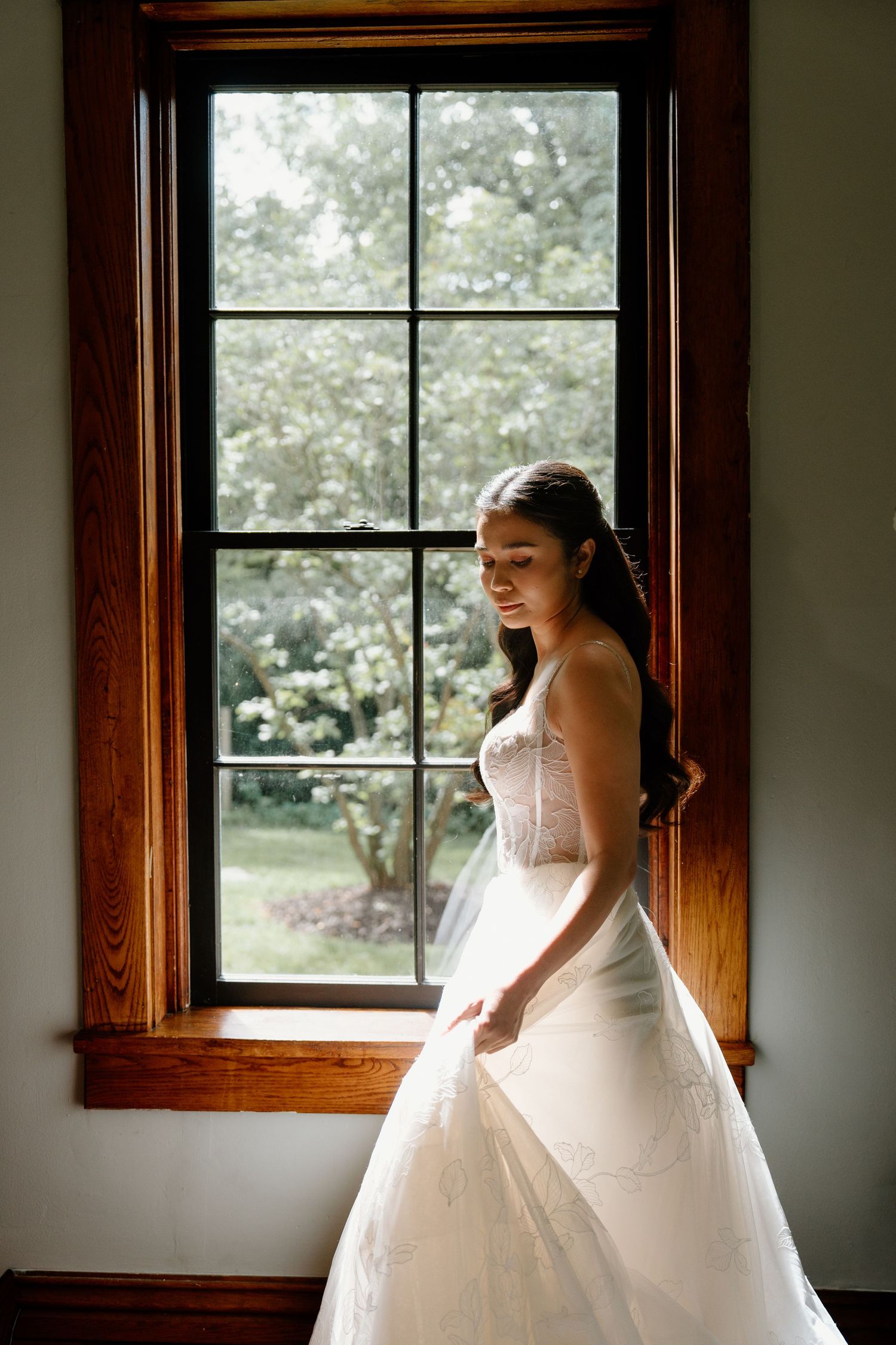 A bride in a white wedding dress stands by a window in a rustic room with natural light streaming in.