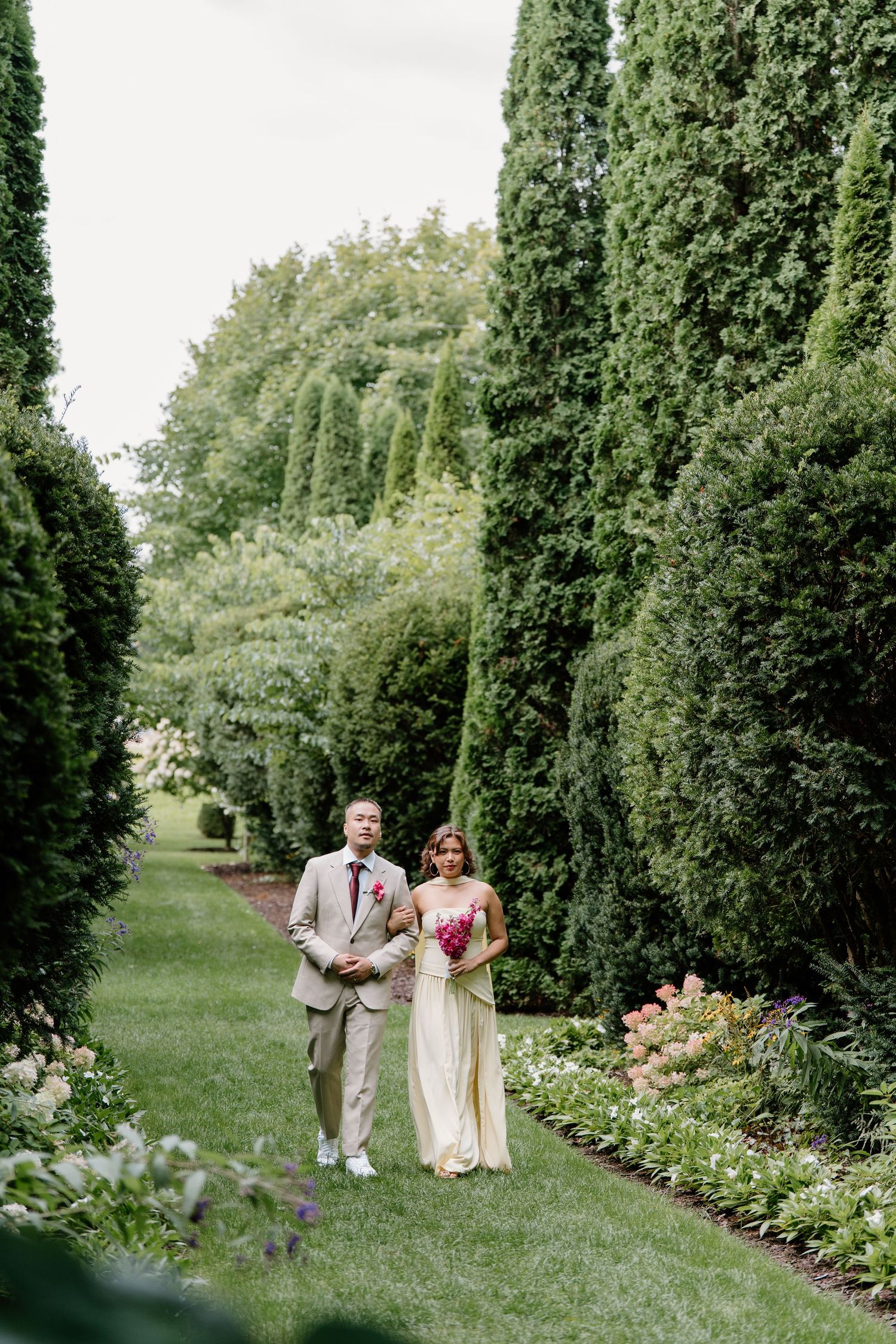 A couple walks through a lush garden path lined with tall evergreen trees and flowering plants.