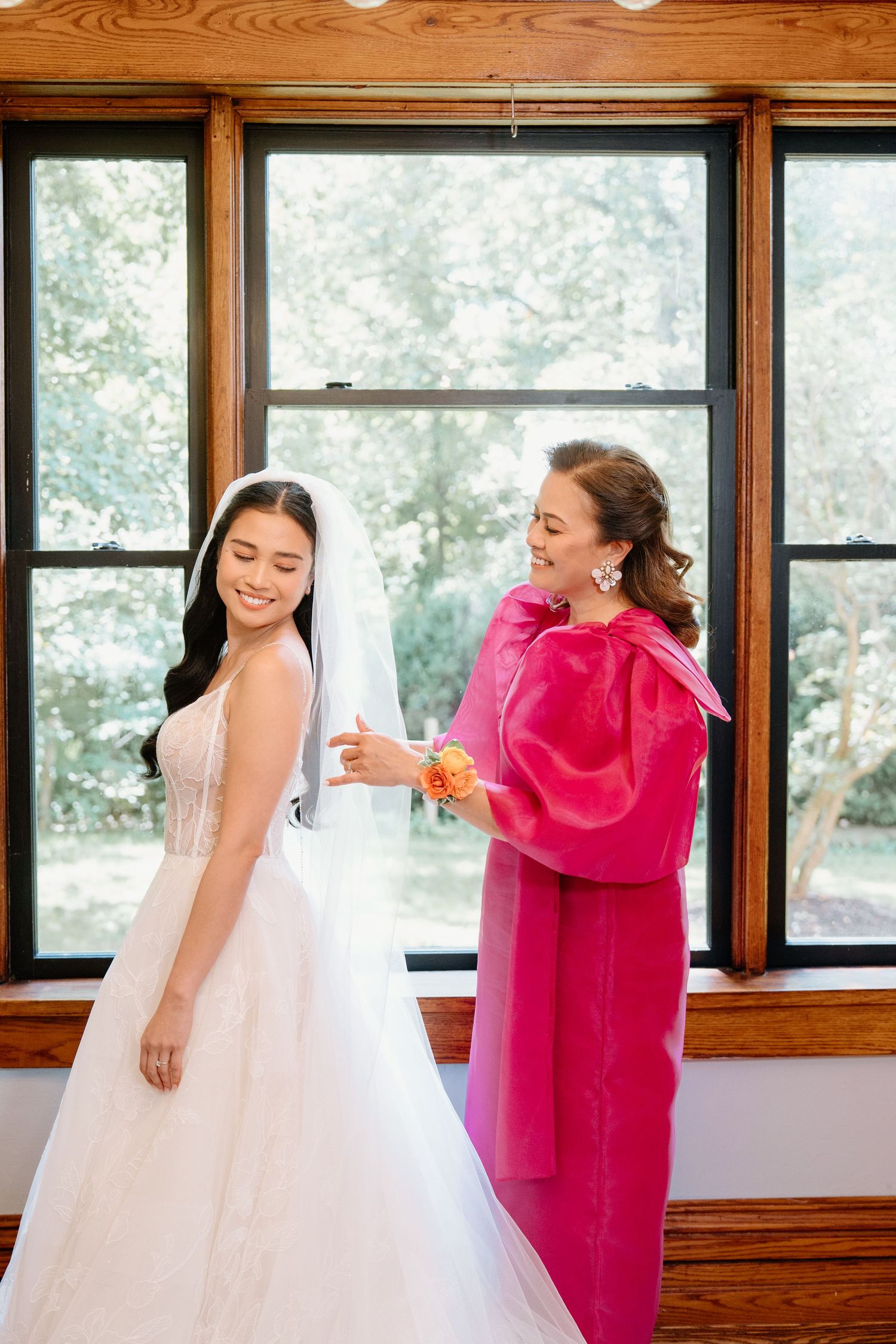 Two people share a joyful moment in a room with wooden walls and windows, one wearing white and one in bright pink.