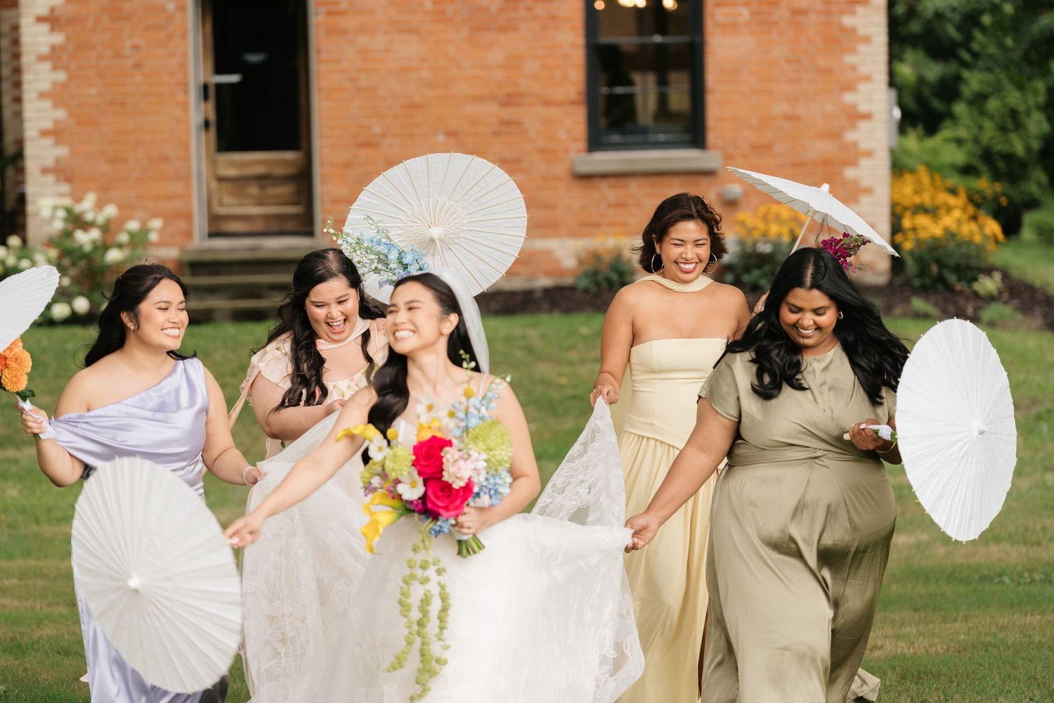 Wedding party members in olive green dresses hold white parasols while posing on a sunny lawn with brick building behind them.