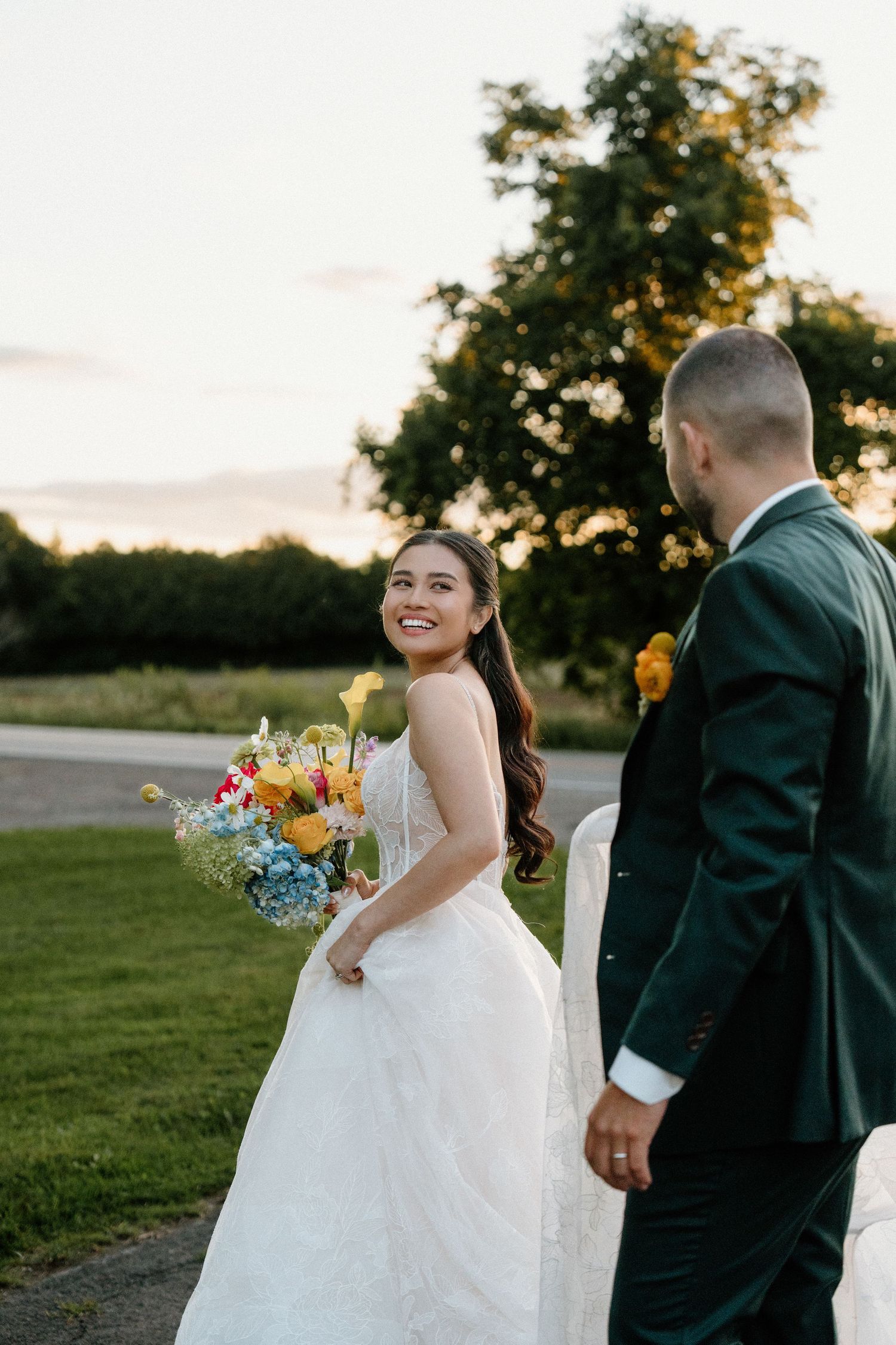 Newlyweds walk together on grassy path at sunset with colorful bouquet.
