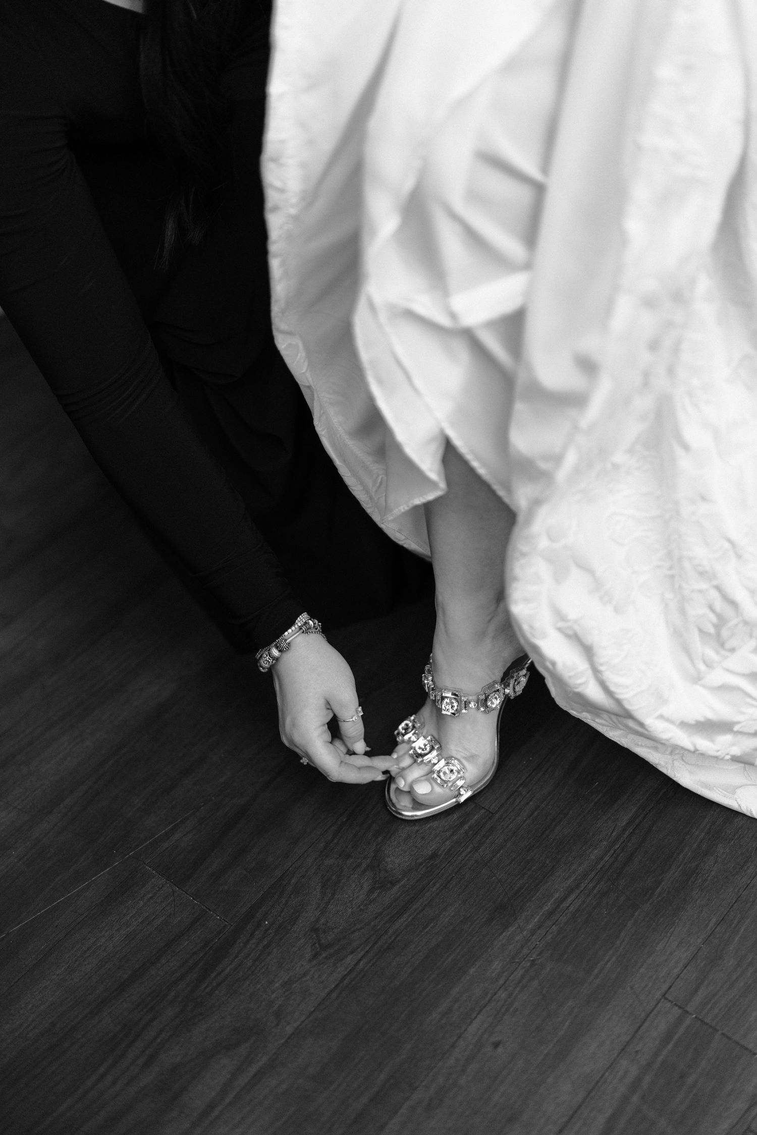 Close-up view of elegant bridal shoes with crystal details peeking out from under a white wedding dress on dark hardwood floor.