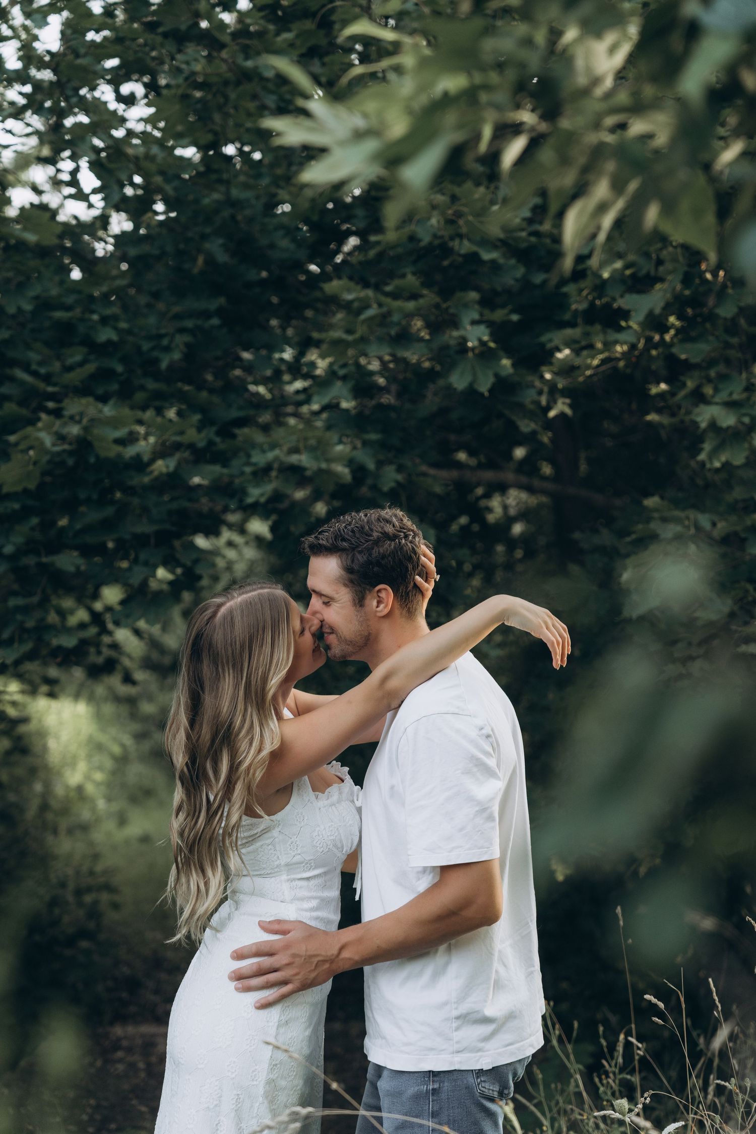 A couple in white outfits embraces romantically in a lush garden setting with green foliage in the background.