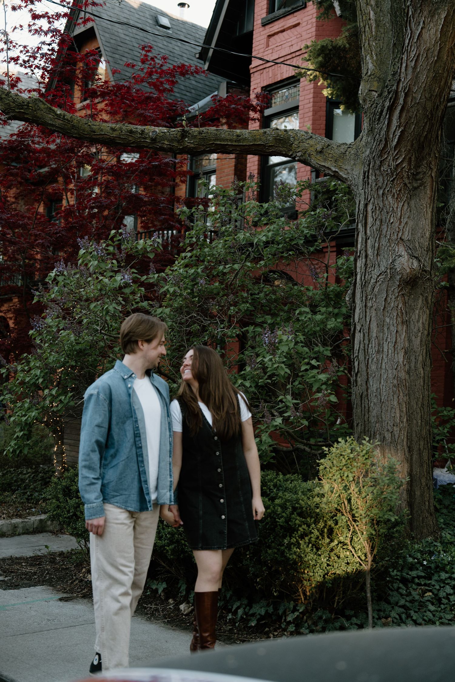 A couple stands close together on a sidewalk near a brick building with red maple trees and lush greenery in autumn.