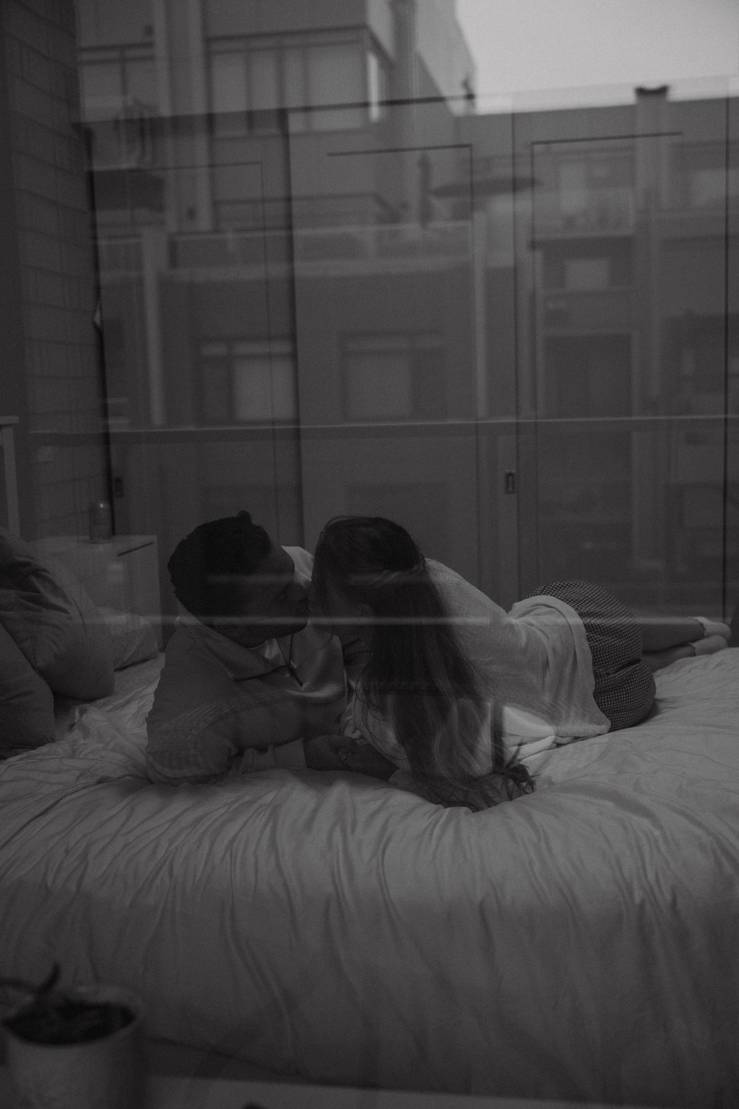 A pair relax together on a bed while looking at something in a moody black and white photograph.