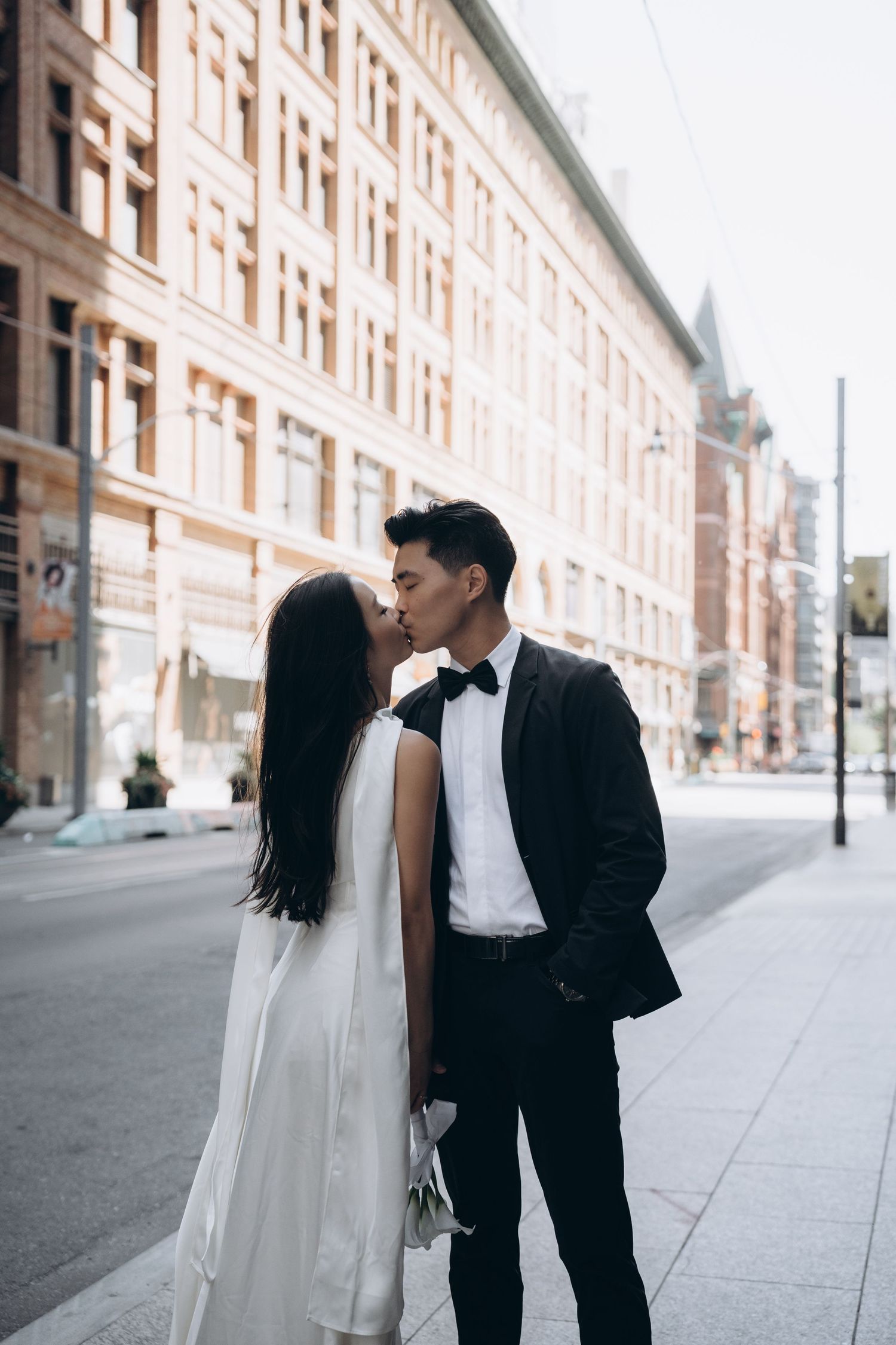 A couple in formal wedding attire share a romantic moment on a city sidewalk with classic architecture in the background.