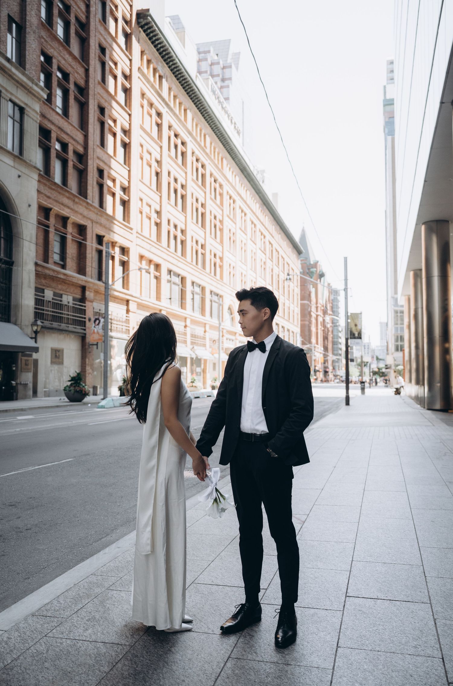 A couple in wedding attire stands on a city sidewalk with historic brick buildings in the background.