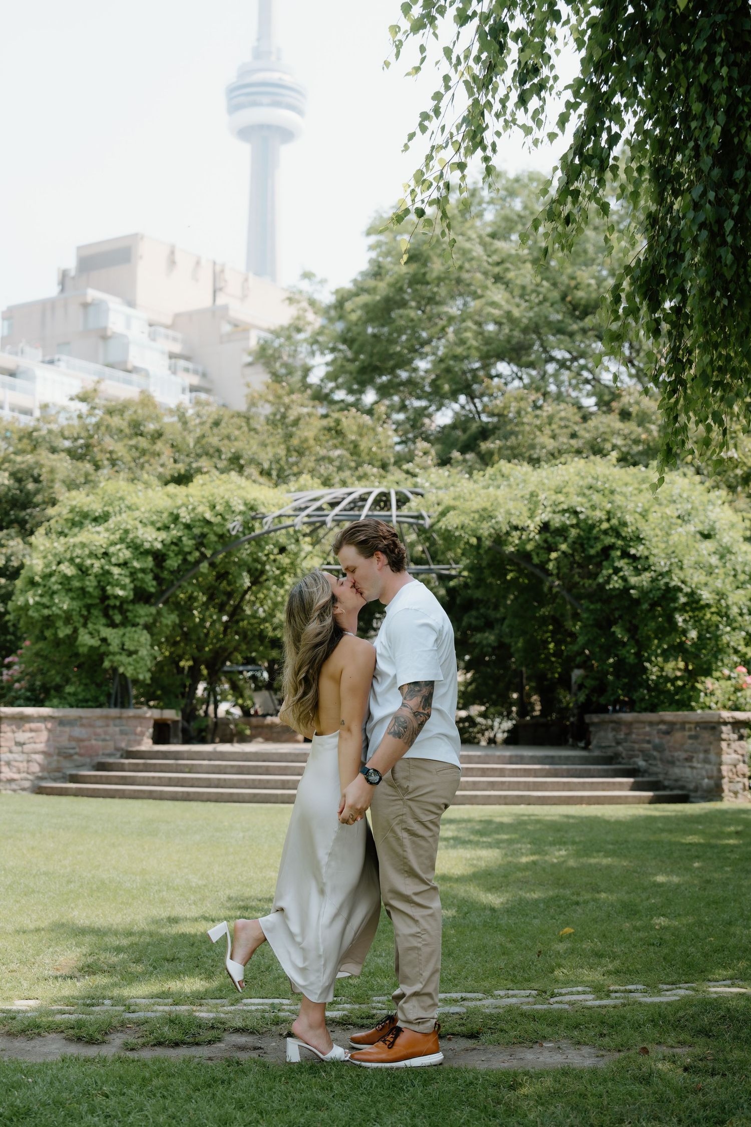 A couple shares a romantic dip kiss in a lush garden setting with an urban backdrop.