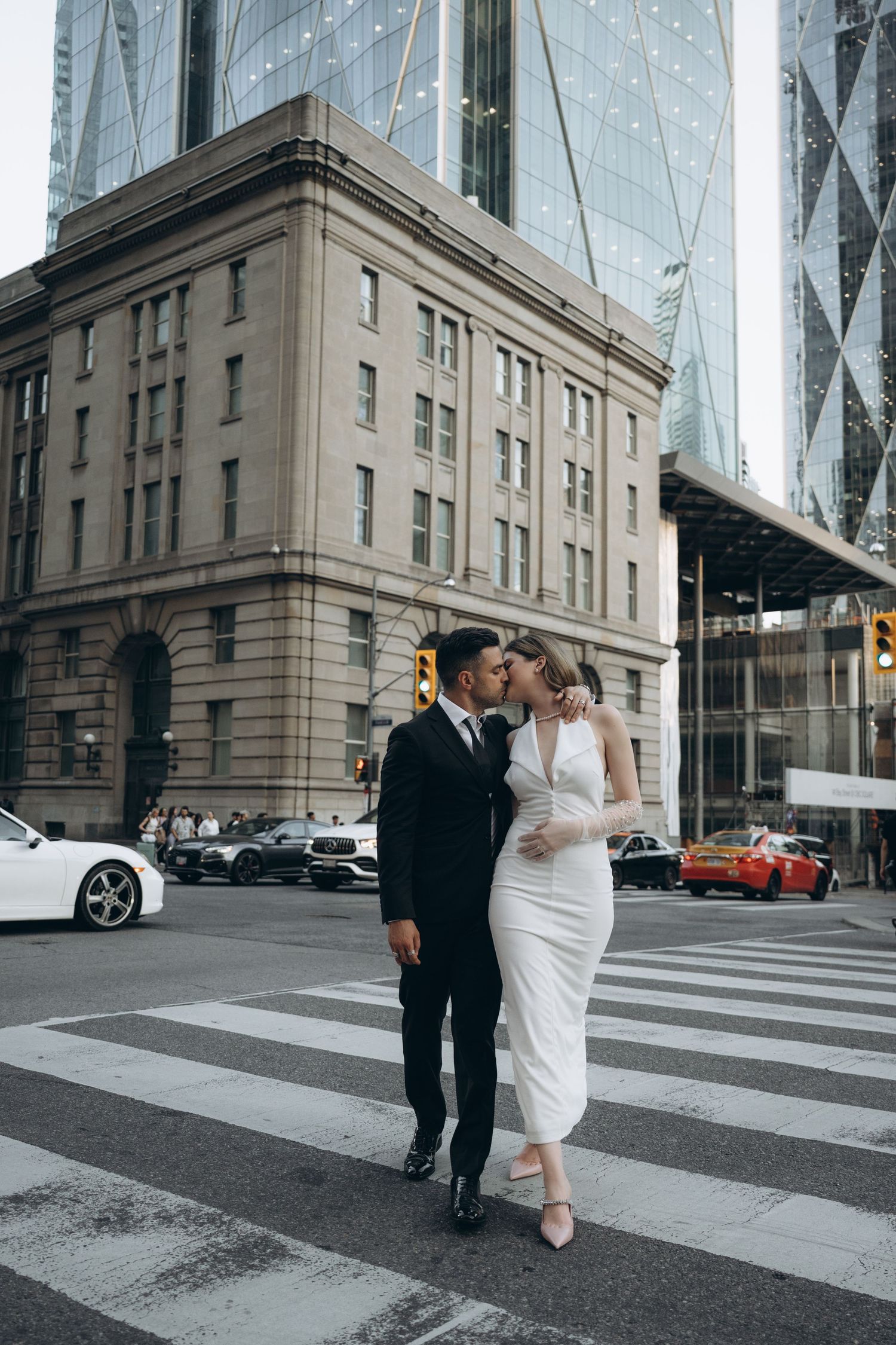 A couple in wedding attire embraces while crossing a city street with modern architecture and classic buildings in background.