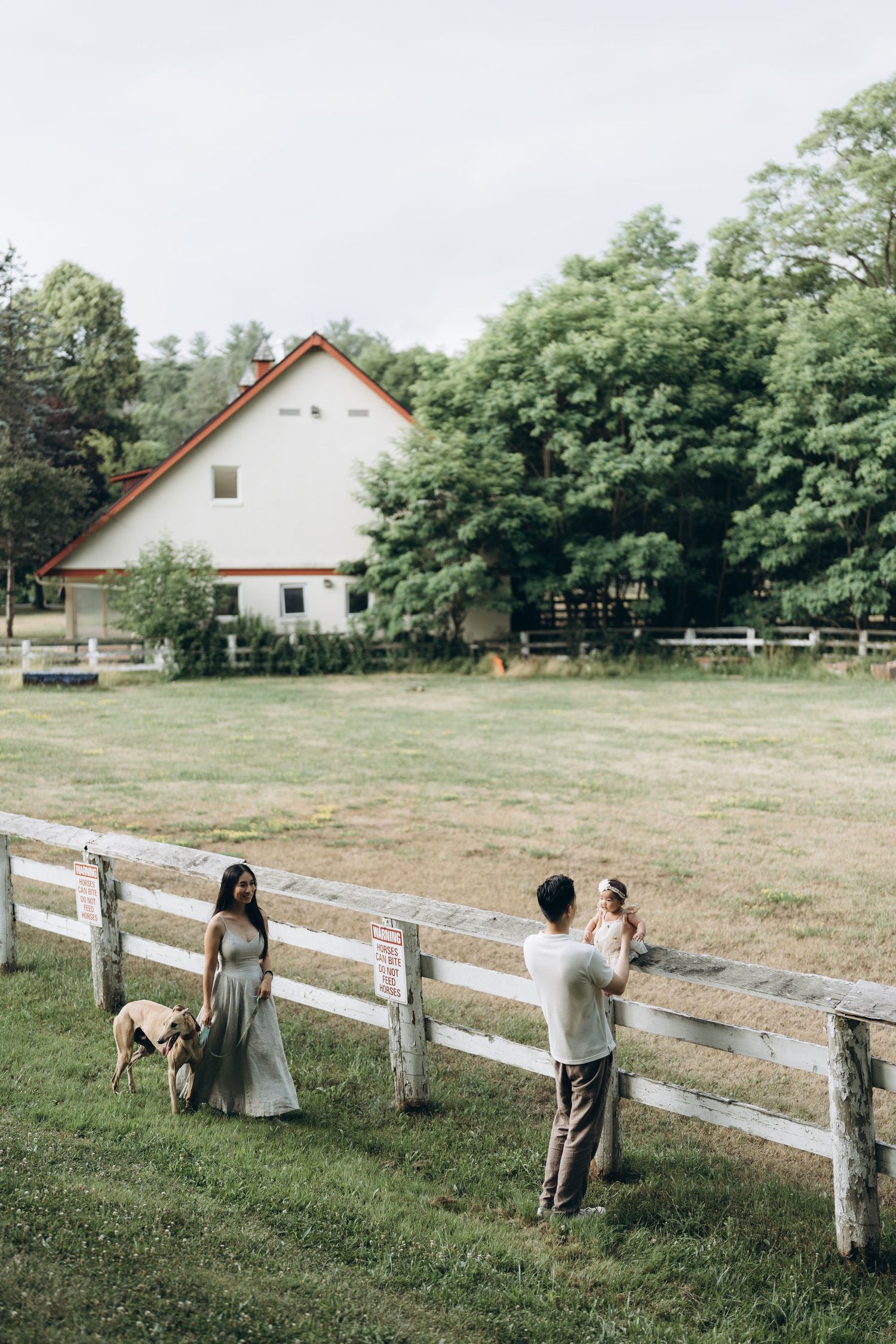 A rustic farmhouse scene with a white fence and people walking along the grass on a sunny day.