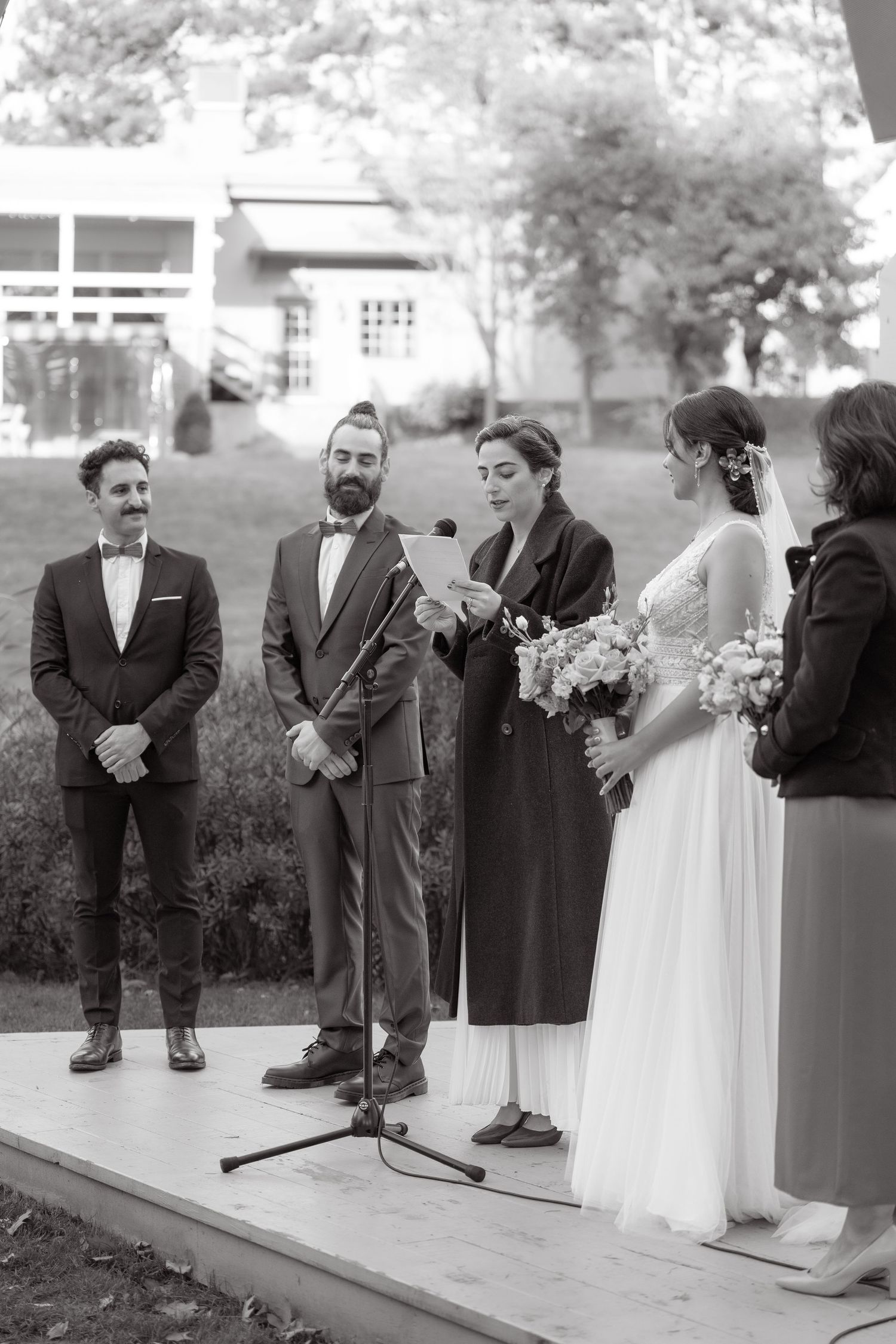 Wedding party stands on a stage during an outdoor ceremony with geometric white backdrop.