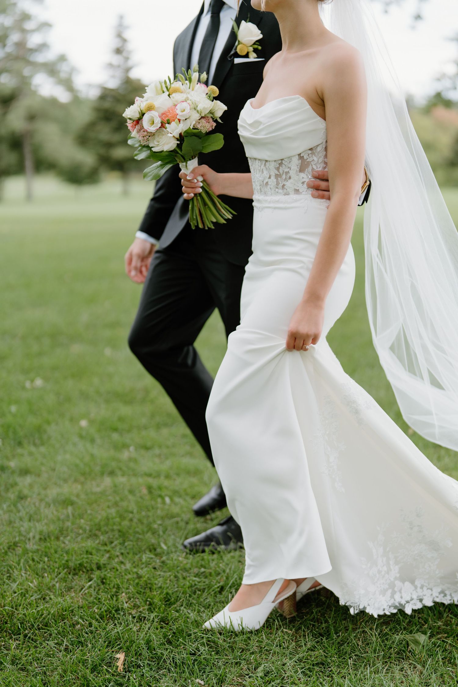 Close-up of bride and groom's feet walking on grass with flowing white wedding dress and veil trailing behind.
