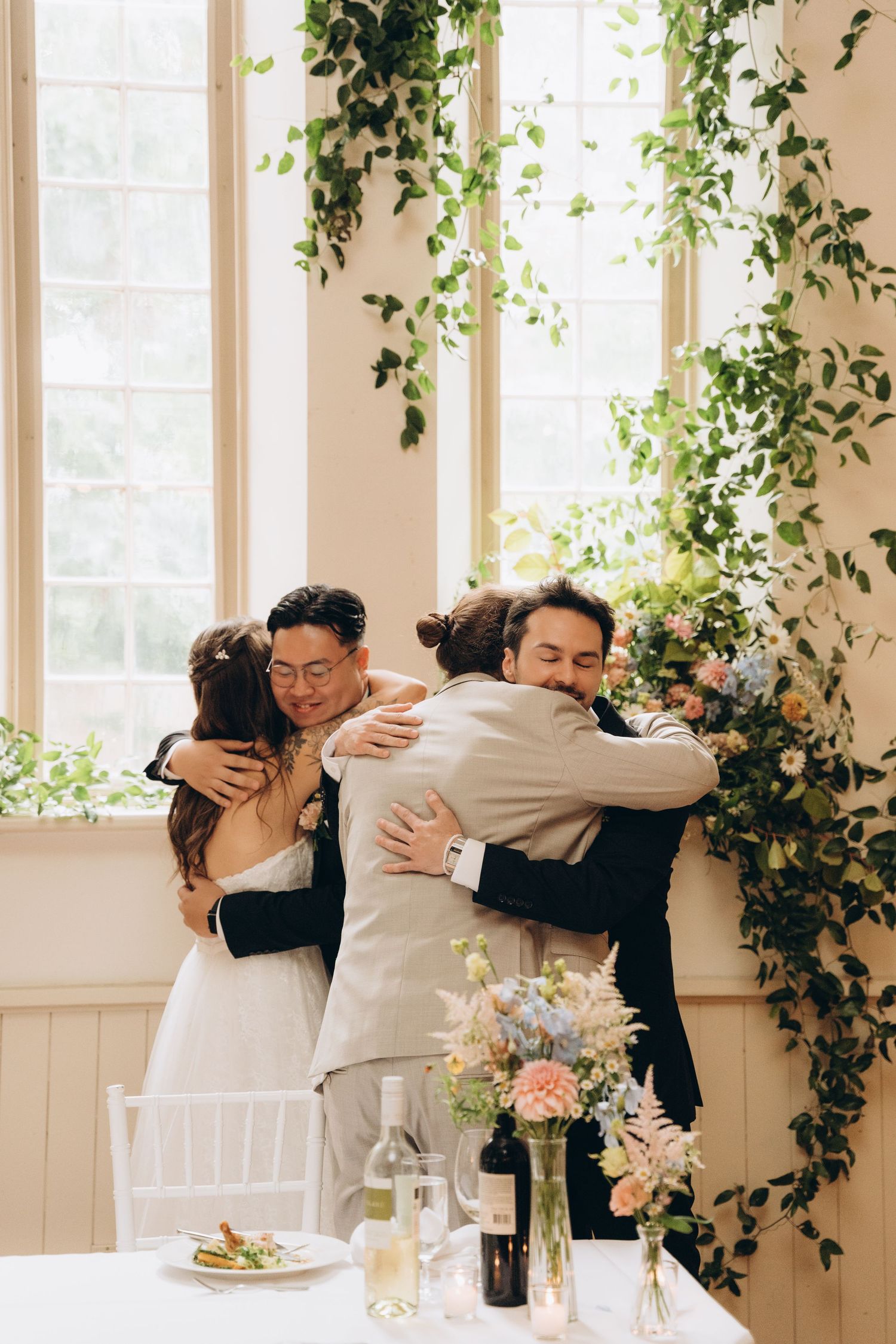 Wedding guests share emotional embraces in a bright room decorated with climbing vines and floral arrangements.