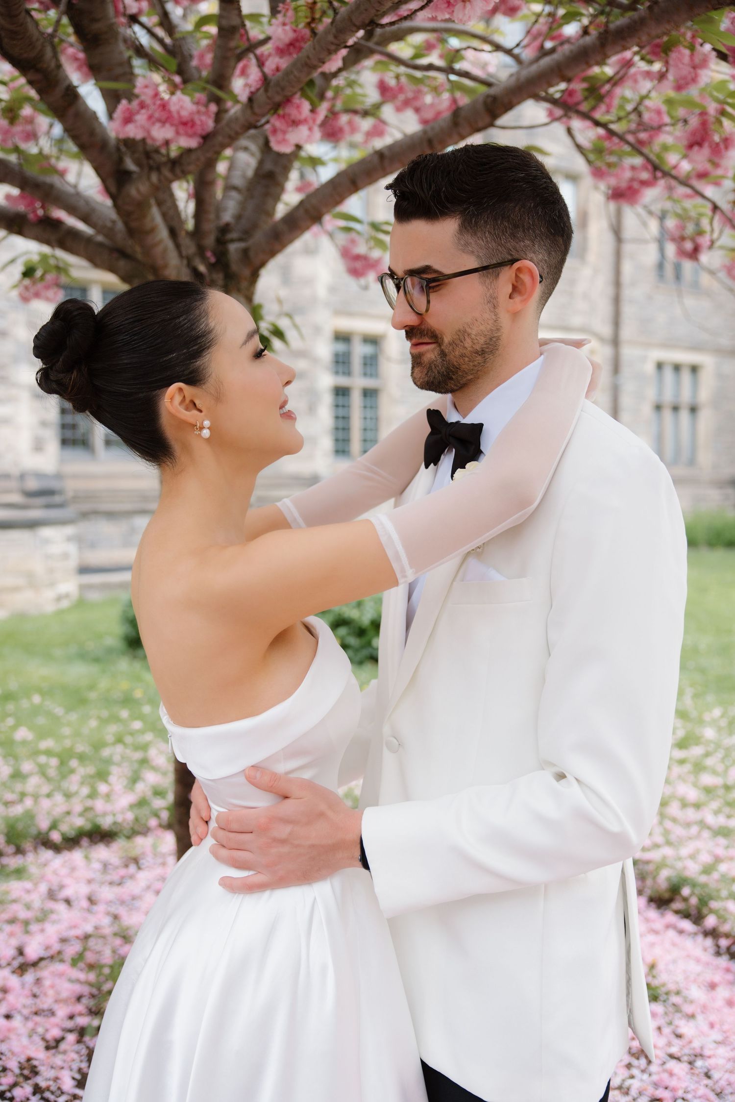 A couple in wedding attire embraces under blooming cherry blossoms on a spring day.