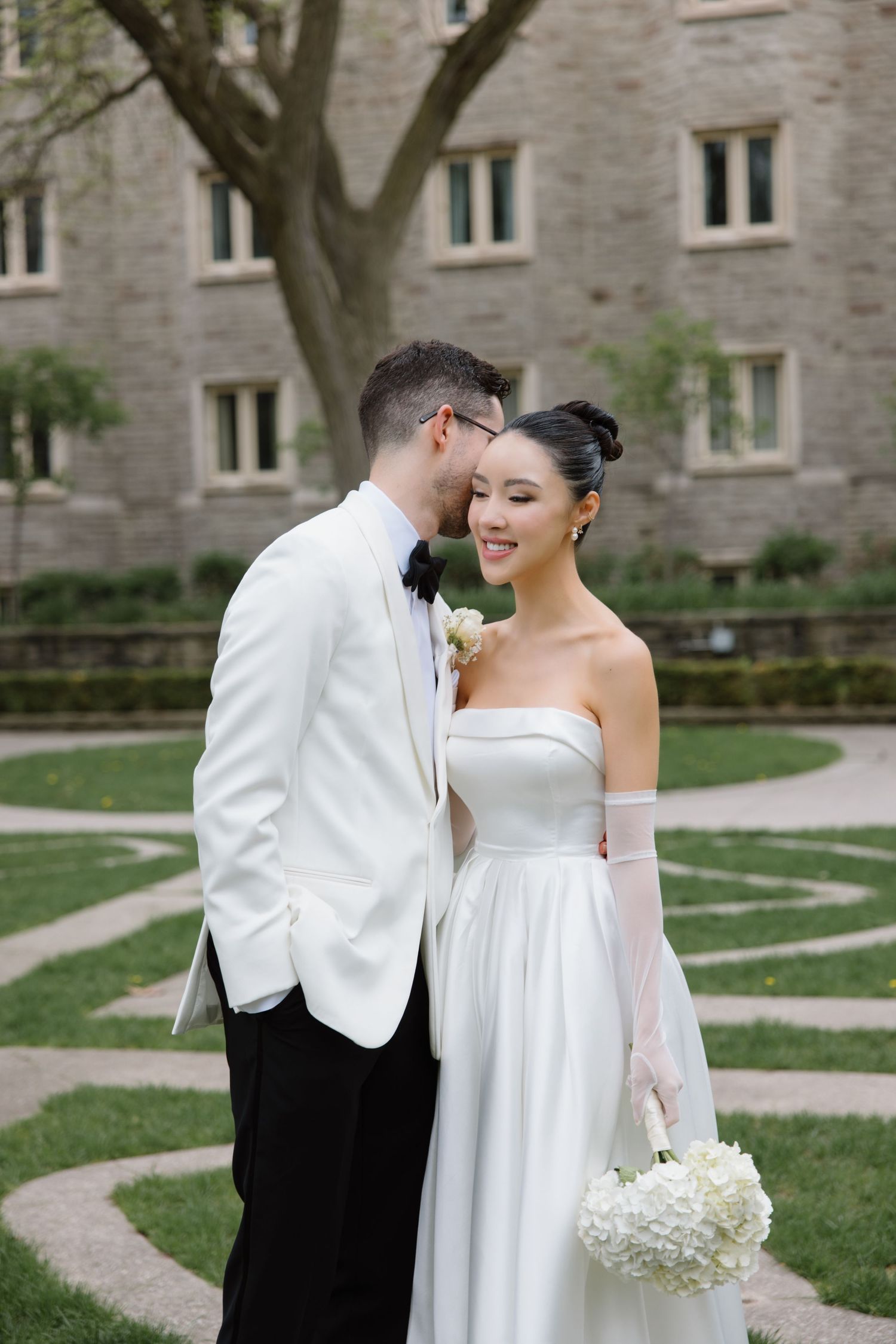A bride in a strapless white gown holds white flowers while sharing an intimate moment outside a stone building.