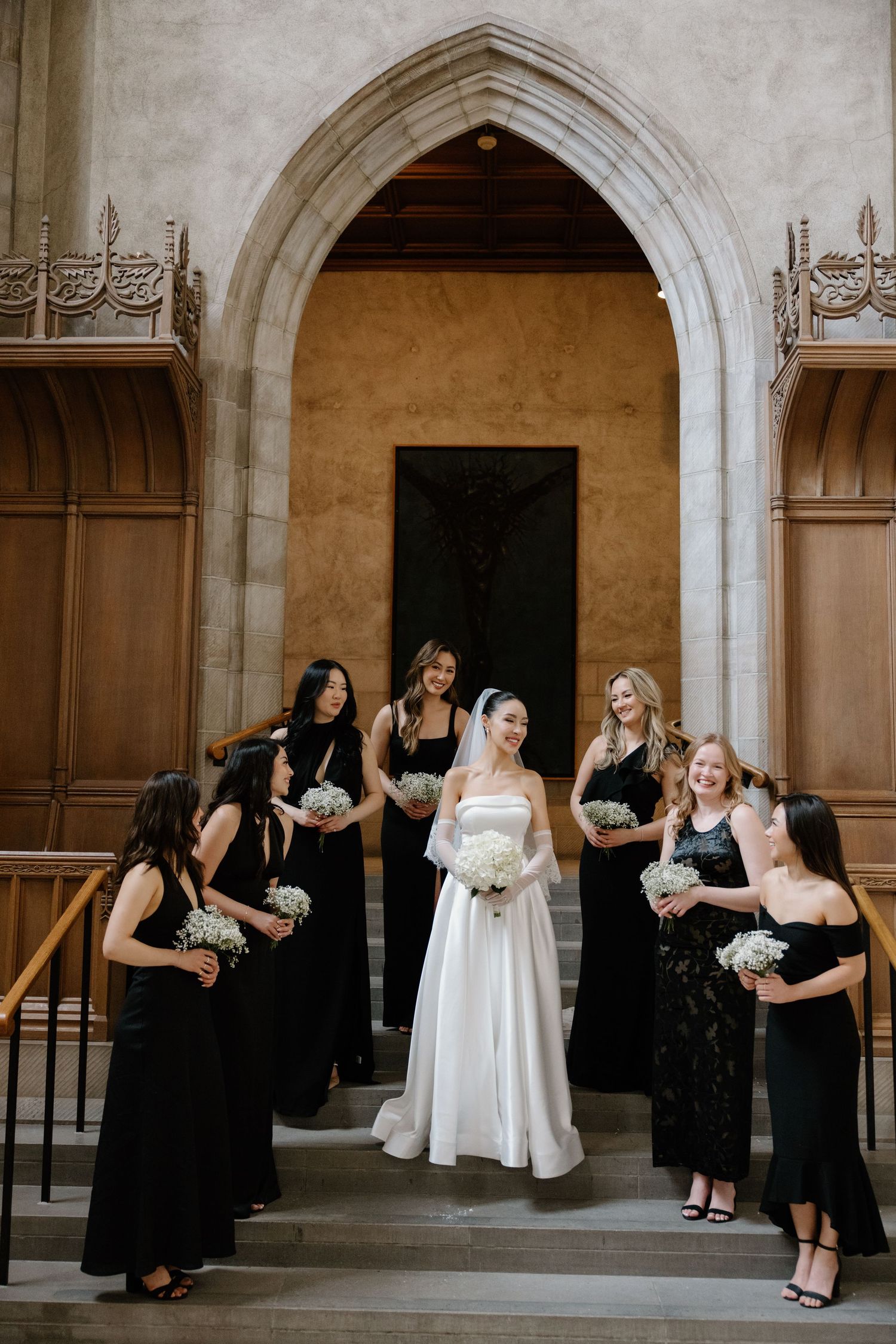 A bride in white gown stands with bridesmaids wearing black dresses on church steps near an arched doorway.