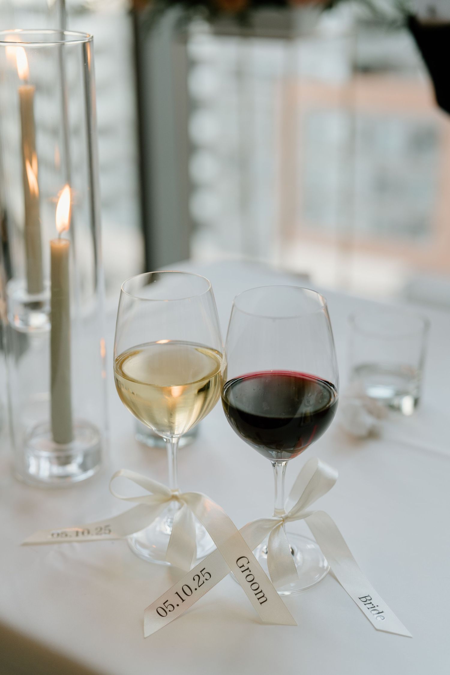 Elegant wine glasses decorated with white ribbons containing red and white wine sit on a white tablecloth with candles.